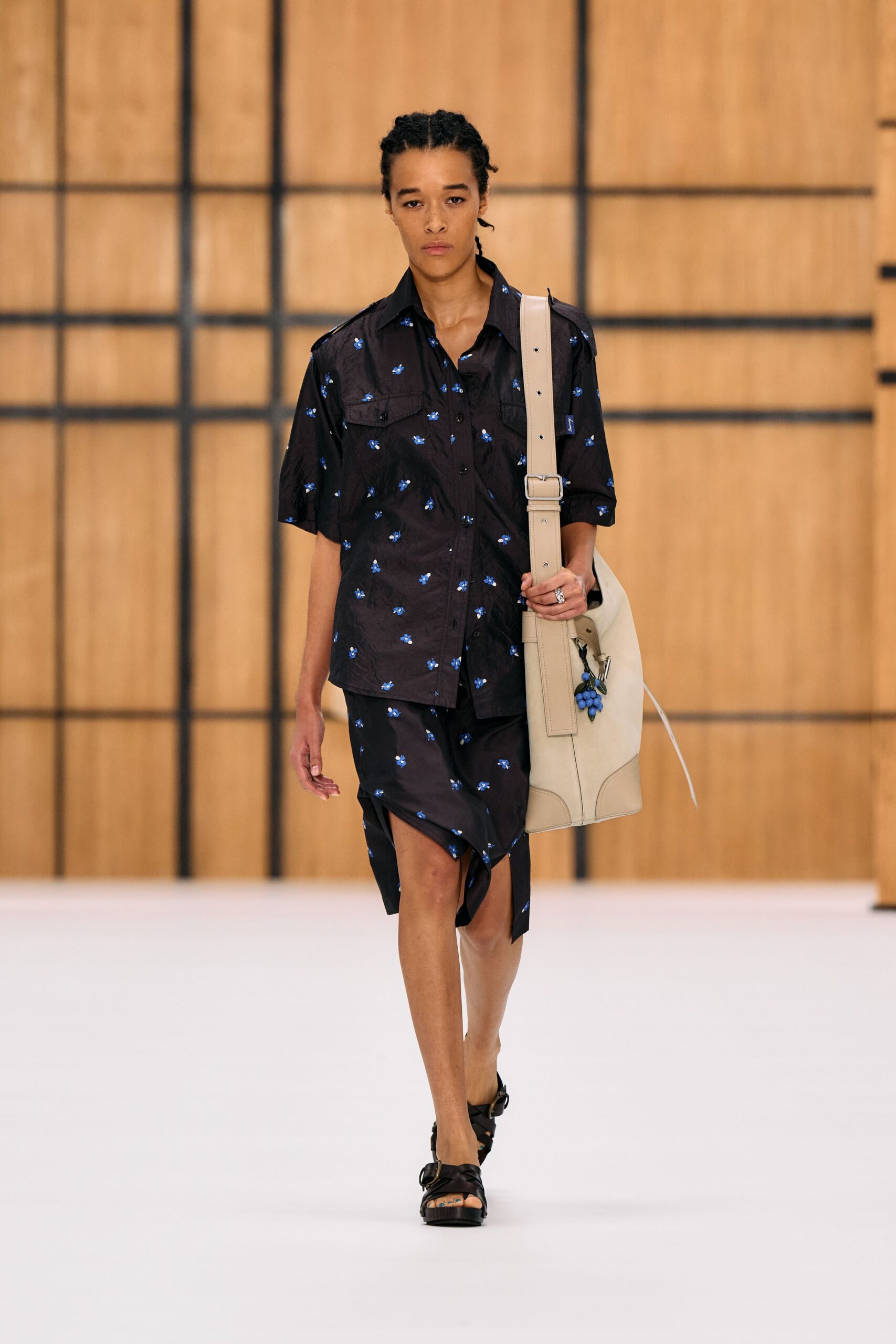 Black camp shirt scattered with tiny blue floral sprigs. Matching wrap skirt, hem kicked into a sharp front point. Oversized chalk leather bag with a bunch of blue grapes clipped on. Black crossover sandals with thick buckled straps. Walking back from the greengrocer with one useful thing and one absurd one.