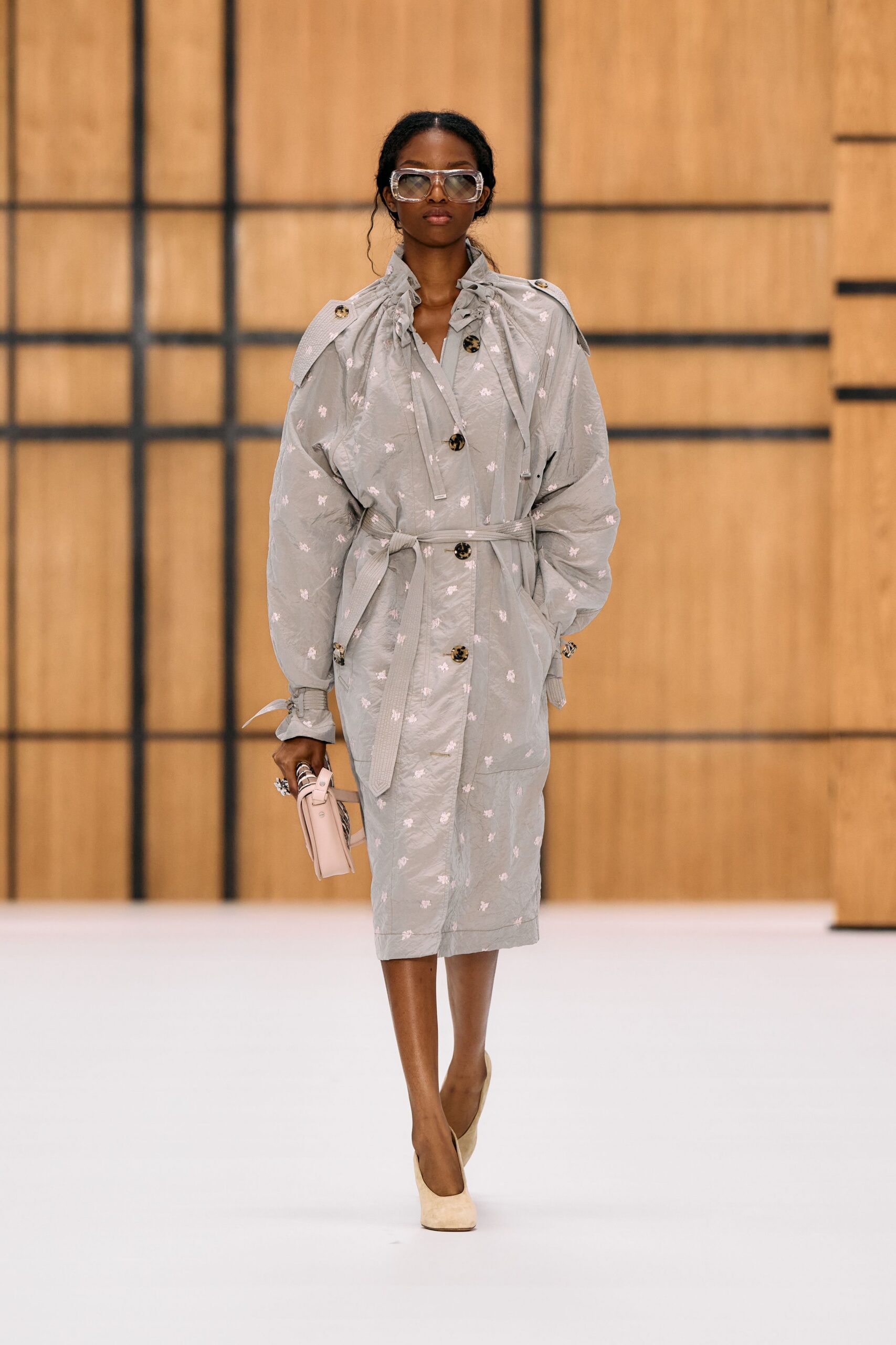 Clear oversized goggles with thick translucent rims. Mist-grey crinkled coat, tiny white motifs, drawstring collar bunched high. Self belt tied loose across the waist. Blush top-handle bag with striped side gusset. Cream suede pumps with a squared toe. Crossing a museum courtyard just after the rain stopped.