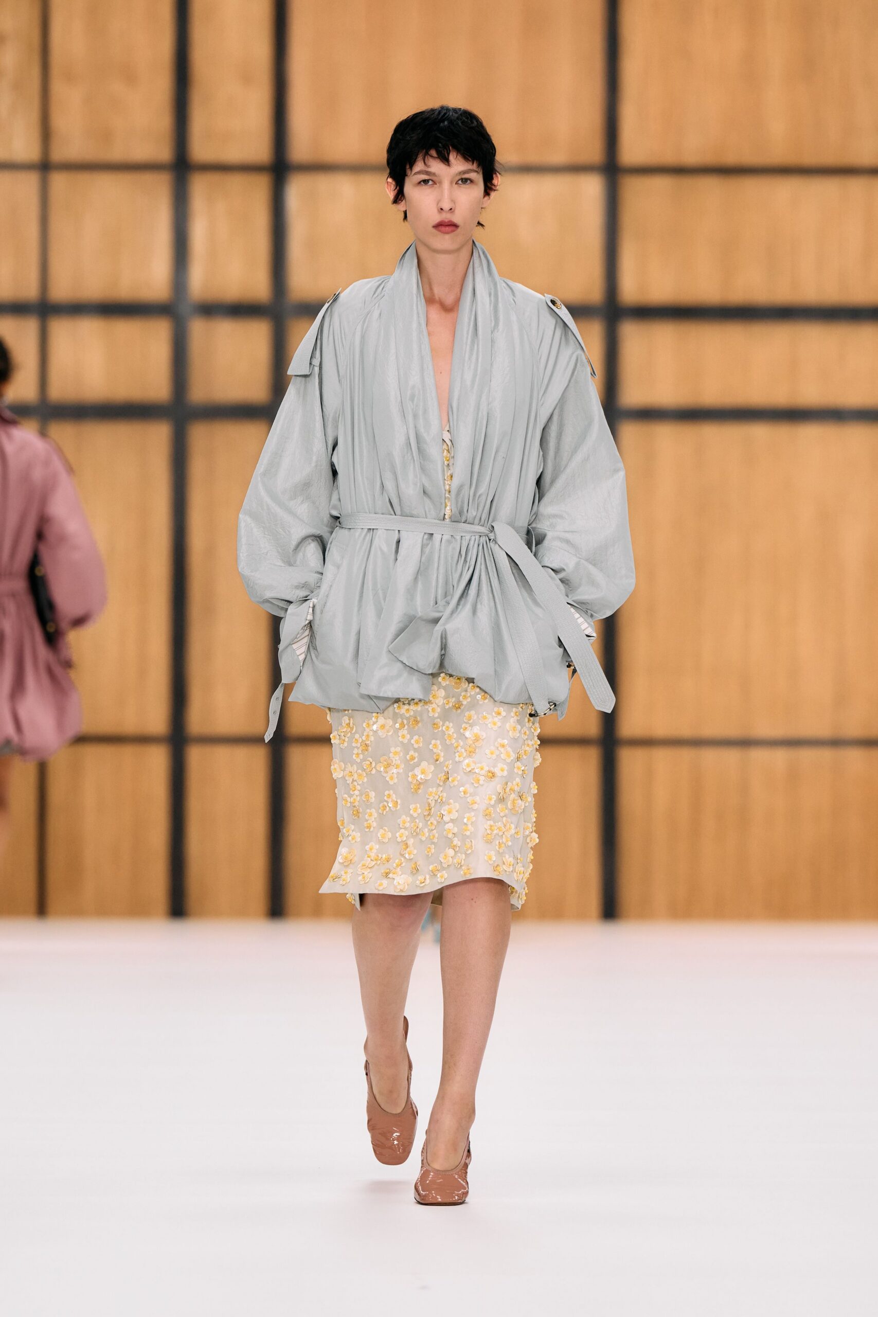Powder-blue crinkled wrap jacket, shawl front collapsing into a tied waist. Cream slip dress scattered with raised yellow floral appliques. Nude patent pumps with a squared toe. Crossing a hotel atrium early, florist buckets still sweating by the service door.
