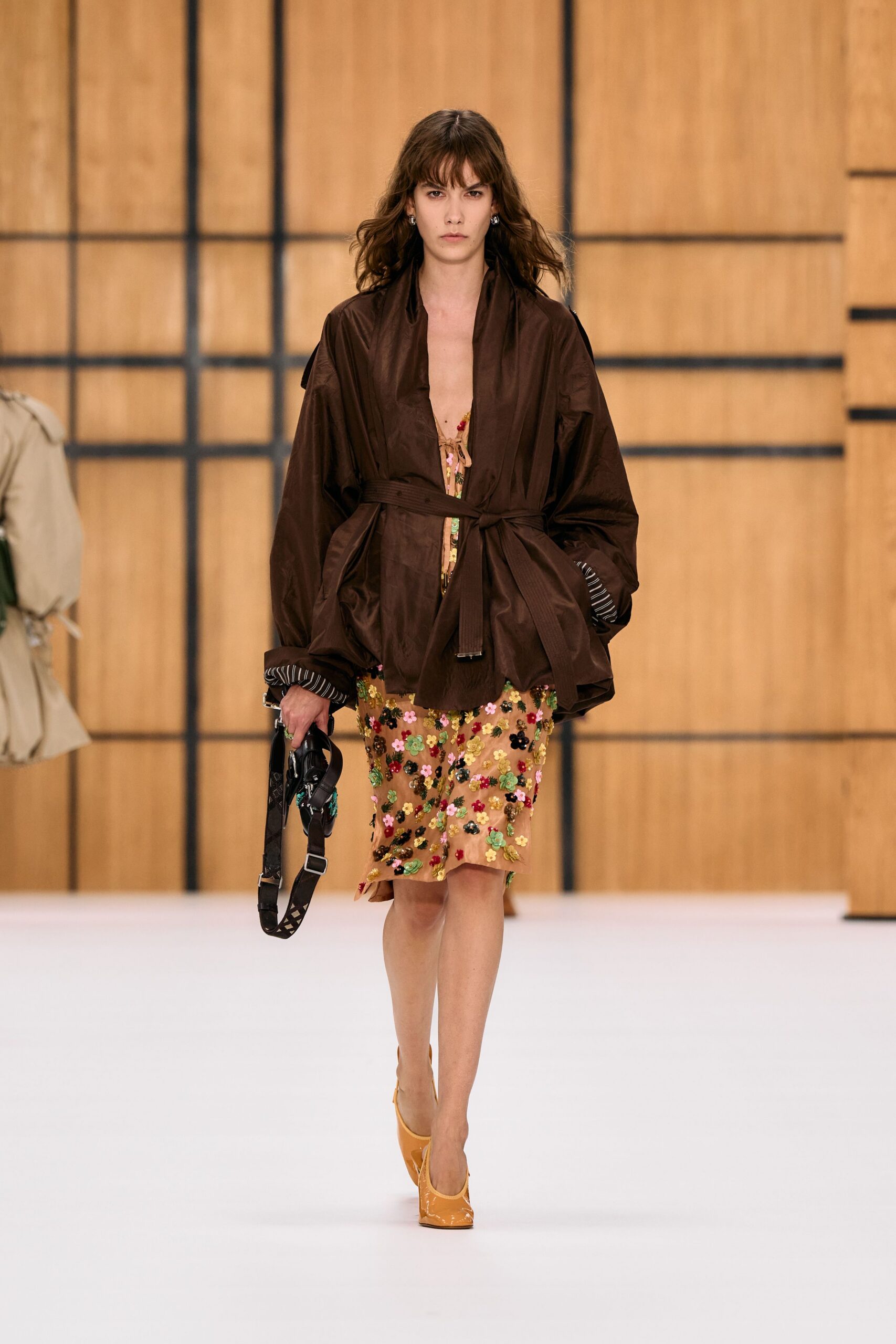 Chocolate paper-nylon wrap jacket, striped cuff lining peeking from balloon sleeves. Tan mesh slip dress thick with tiny sequined flowers, hem tilting uneven. Black shoulder bag dangling by its webbing strap. Butterscotch patent pumps with squared vamps. Crossing a hotel atrium after the wedding breakfast ran long.