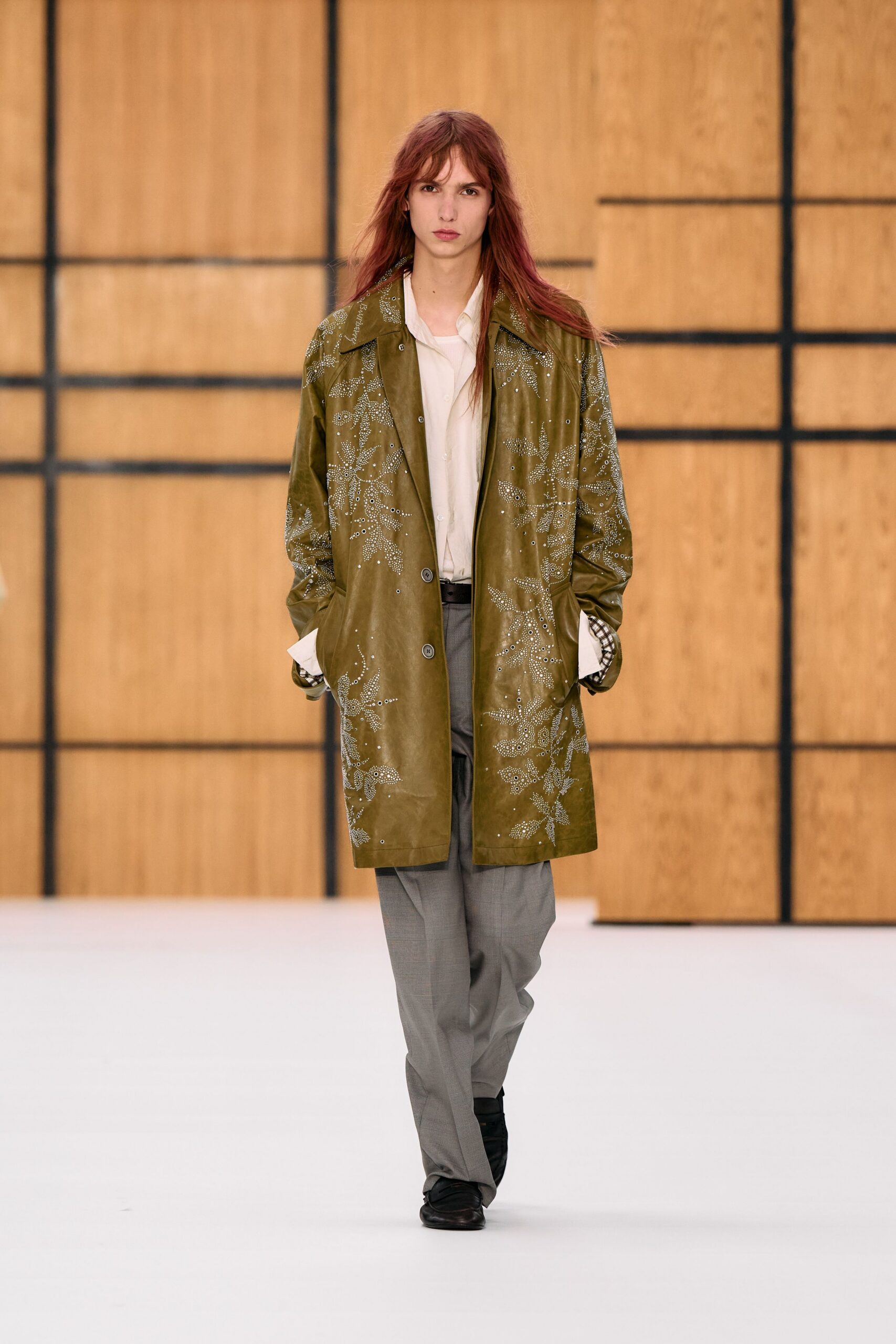 Olive leather car coat punched with silver floral studs and eyelets. Soft ivory shirt, long cuffs falling past the coat sleeves. Grey micro-check trousers with a loose straight break. Black square-toe shoes. Early train north, station coffee cooling through your gloves.