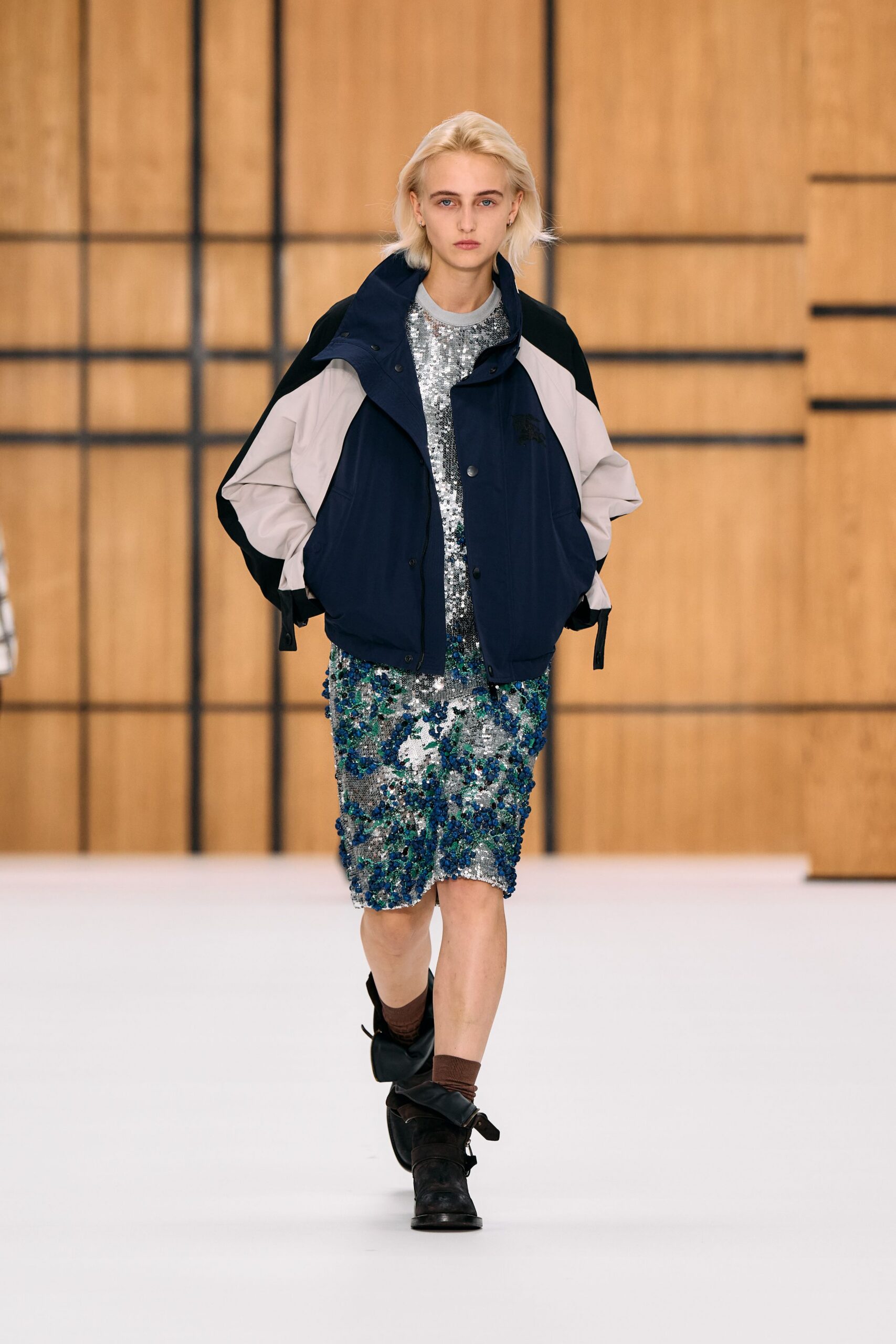Navy and chalk color-block windbreaker with a high snap stand collar. Silver sequined shift dress crusted with cobalt and emerald paillettes. Brown ribbed socks slouching above the ankle. Black suede shoes tied with oversized floppy bows. Walking from the gym into a hotel lobby still full of wedding confetti.