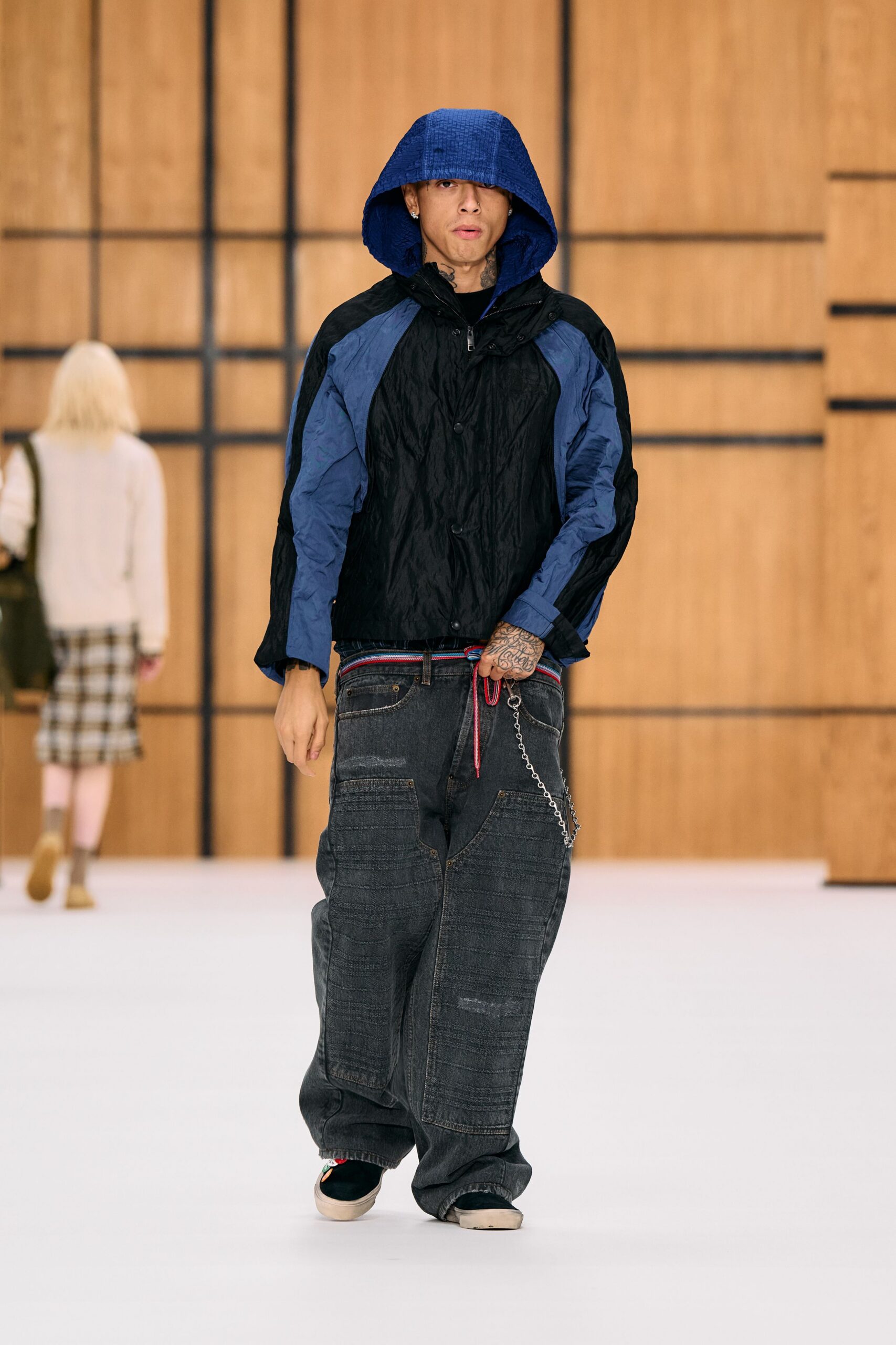 Black crinkled shell jacket with slate raglan panels and a deep cobalt hood. Red and blue cord looped at the waist, ends hanging long. Faded black wide jeans with ribbed knee panels and a wallet chain. Black skate slip-ons with thick off-white rubber foxing. Walking under station eaves after rain, hood still up against the drip.