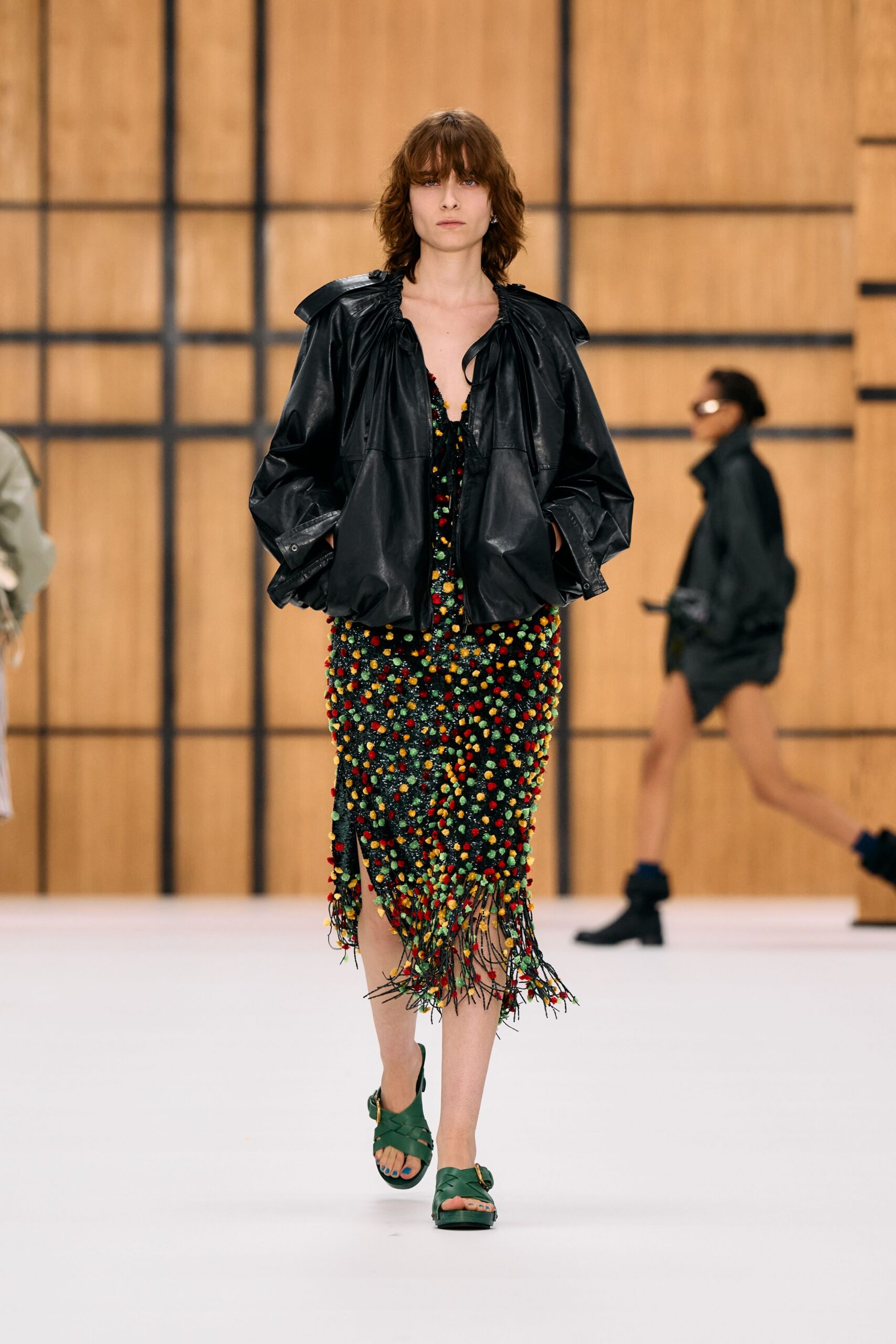 Black leather blouson with gathered neckline and puffed, folded shoulders. Black net midi crusted with tiny red, yellow and green tufts. Long black threads swishing loose below the calf. Green woven leather platform sandals with a gold side buckle. Walking through a conservatory after rain, shoes loud on the paving.
