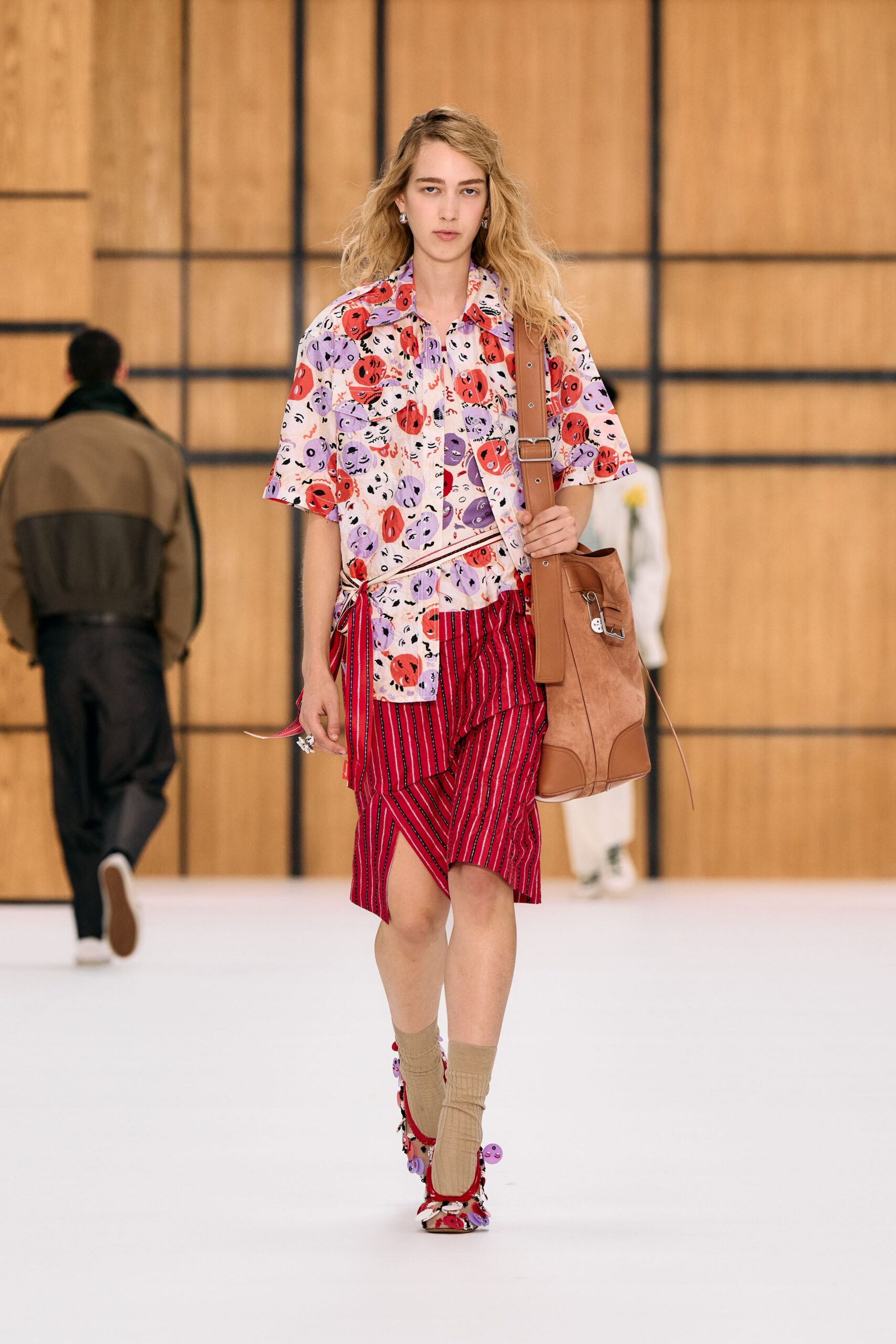 Cream camp shirt printed with red and lilac cartoon faces. Red striped wrap skirt, one panel skewed open at the knee. Tan suede shoulder bag with a wide buckled strap. Beige ribbed socks scrunched at the ankle. Print-mixed heels dotted with lilac disc charms. Walking into a hotel breakfast still wearing last night's wristband.