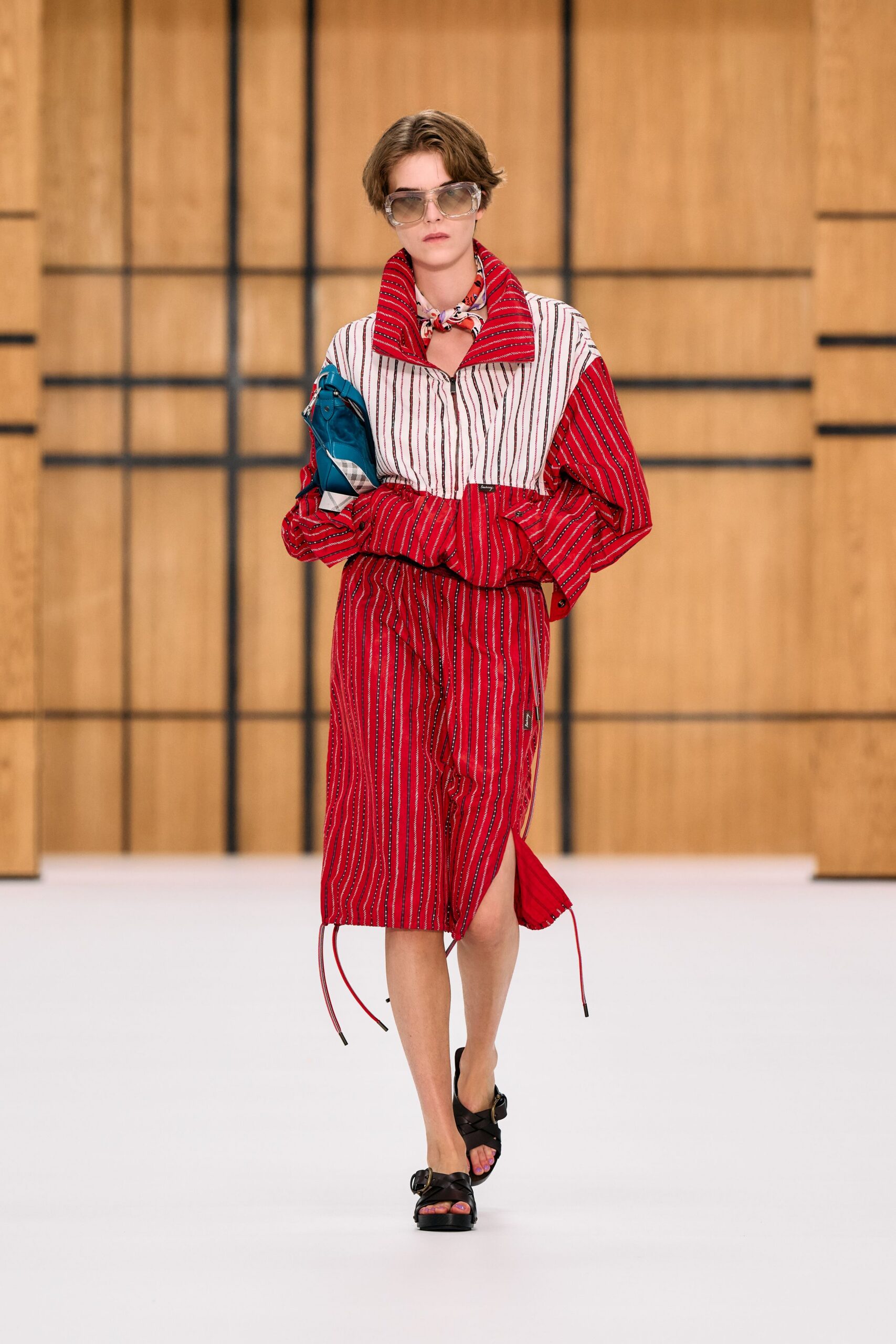 Oversized smoke-tinted visor frames. Printed silk neckerchief knotted close under the chin. Red and white striped zip-front dress, funnel collar flaring wide. Teal nylon pouch tucked under one arm. Black leather sandals with thick crossover straps and brass buckles. Walking from a ferry terminal with salt drying on your neck.