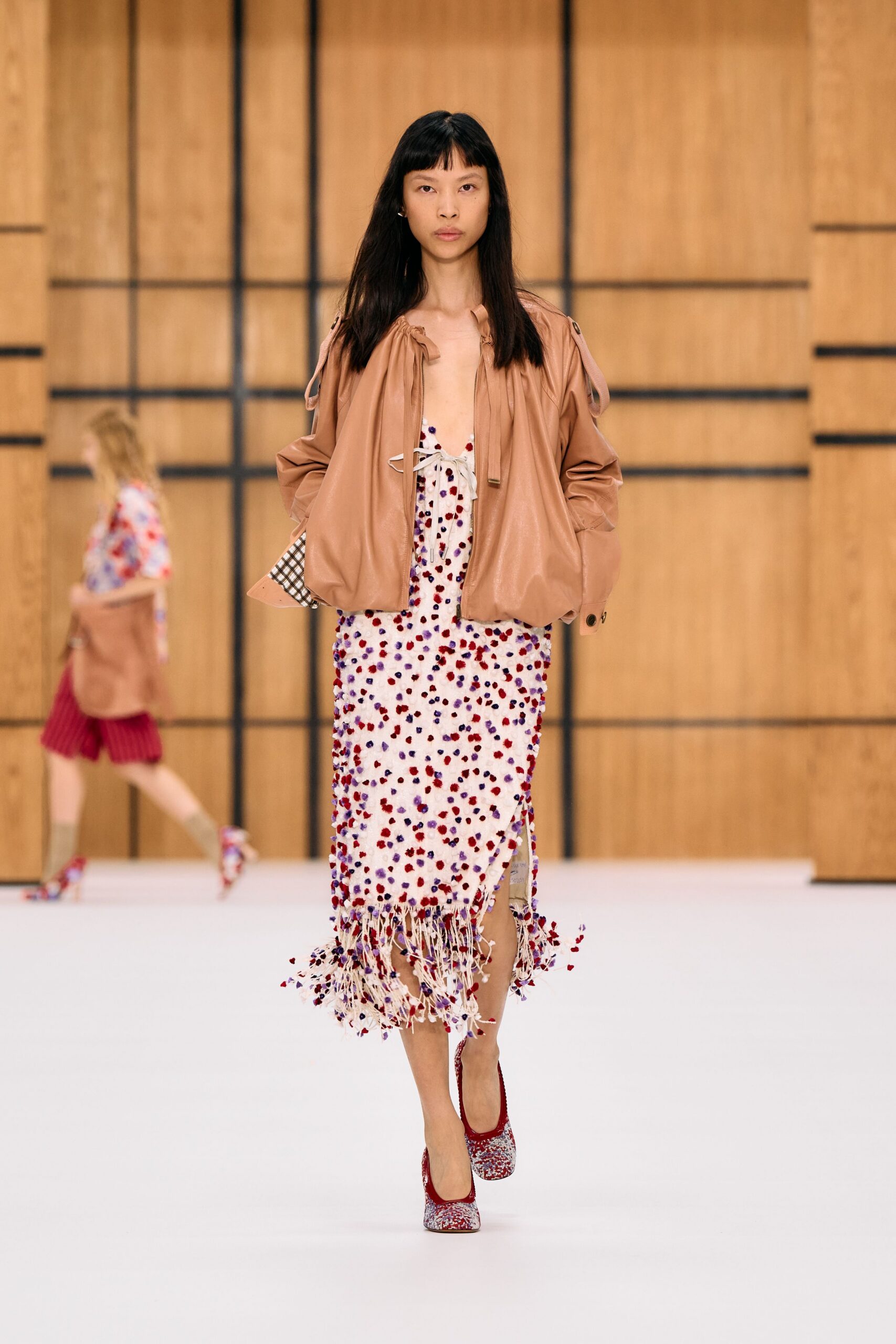 Blush leather blouson, shoulder tabs loose and hem ballooned inward. Ivory open-knit slip dress dotted with tufted red and violet knots. Long cream fringe swinging below the calf, side slit flashing lining. Red-trimmed pumps in dense multicolour bouclé. Walking through a cedar-lined atrium after rain, carrying petals on the hem.