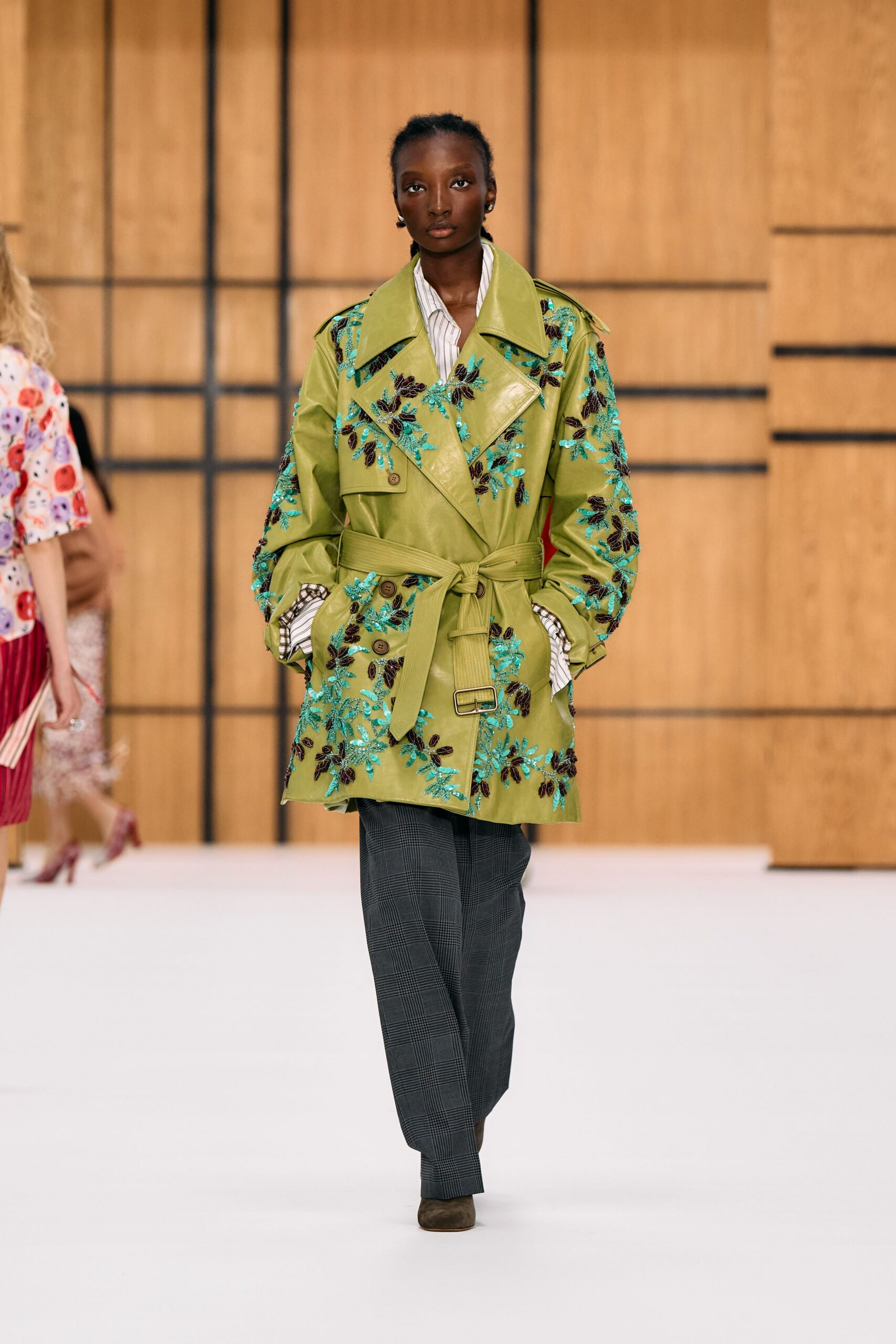 Chartreuse leather trench densely embroidered with turquoise sequins and dark paillettes. White striped shirt, cuffs spilling long past the sleeves. Charcoal glen plaid trousers, cut easy through the leg. Moss suede square-toe ankle boots. Walking into a hotel lobby still carrying rain on the hem.