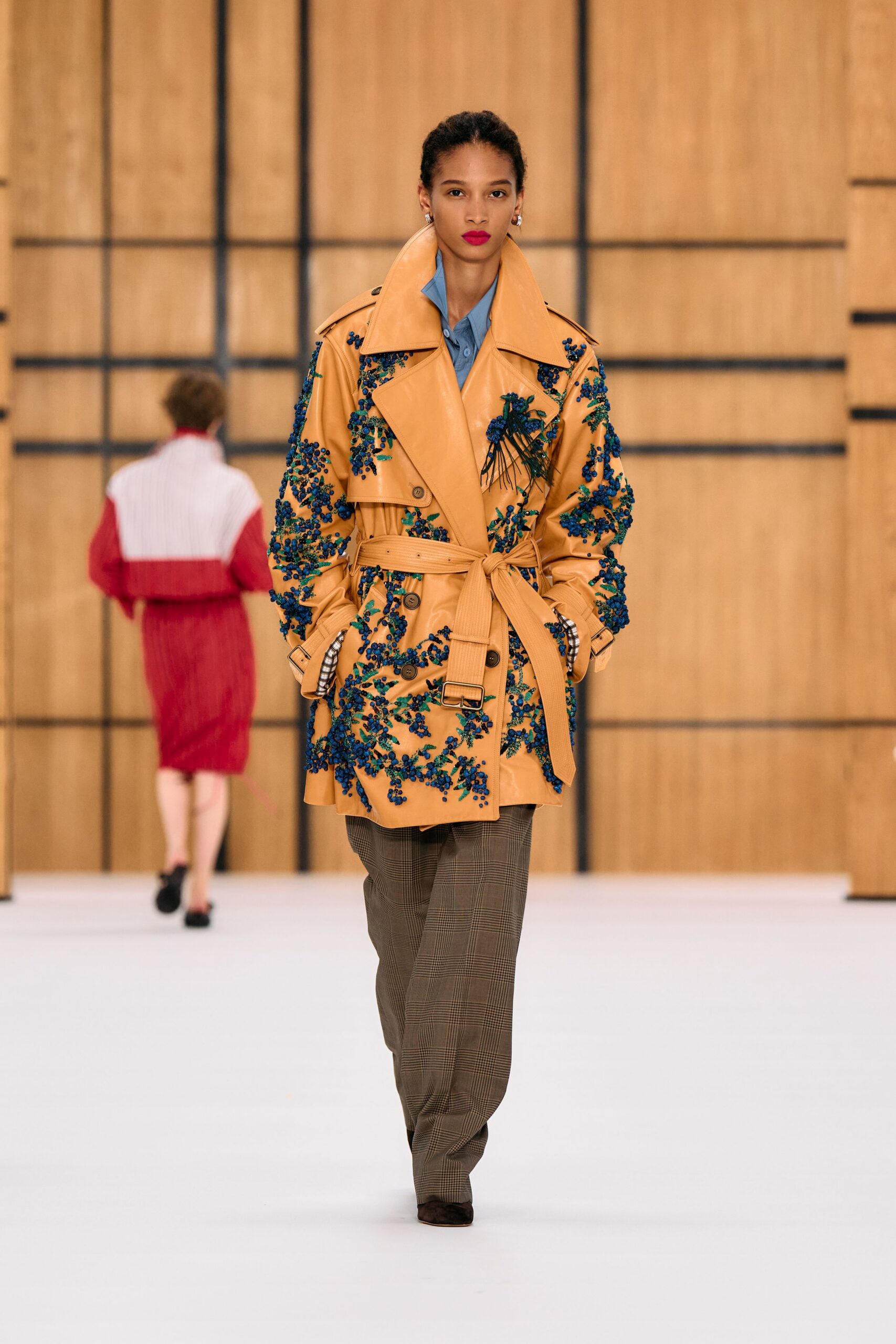 Butterscotch leather trench thick with cobalt beaded sprigs, belt knotted off-center. Powder-blue shirt peeking through the raised lapels. Soft brown glen plaid trousers falling loose over the shoe. Dark suede pointed shoes. Crossing a hotel atrium with flowers still wrapped in wet paper.