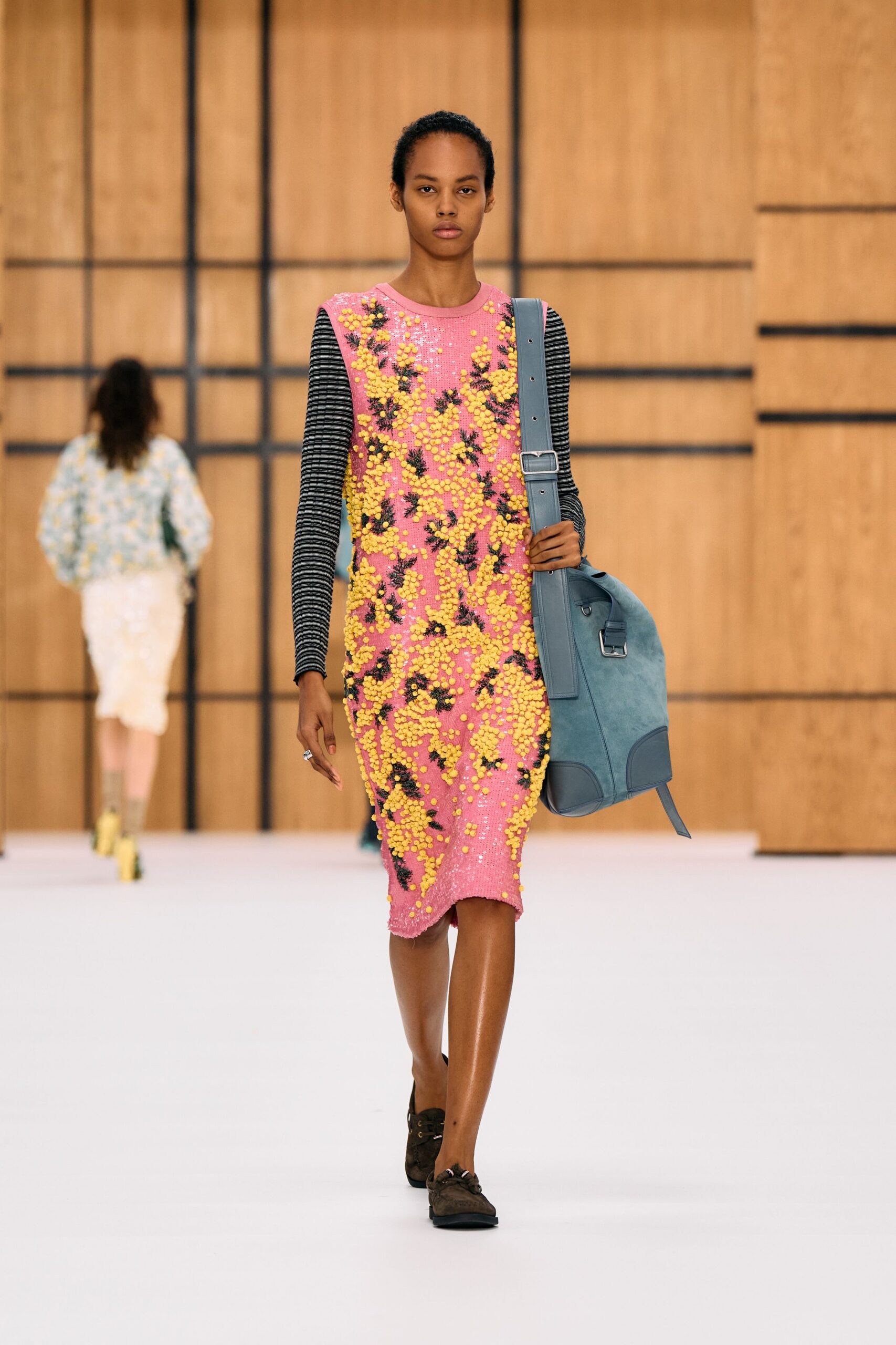 Pink sequined column dress densely appliqued with yellow mimosa clusters. Black-and-white ribbed sleeves extending tight beneath the sleeveless shell. Dusty blue suede carryall with an extra-wide buckled strap. Dark suede lace-up flats with blunt rounded toes. Crossing a ferry terminal with cut flowers and too much to carry.