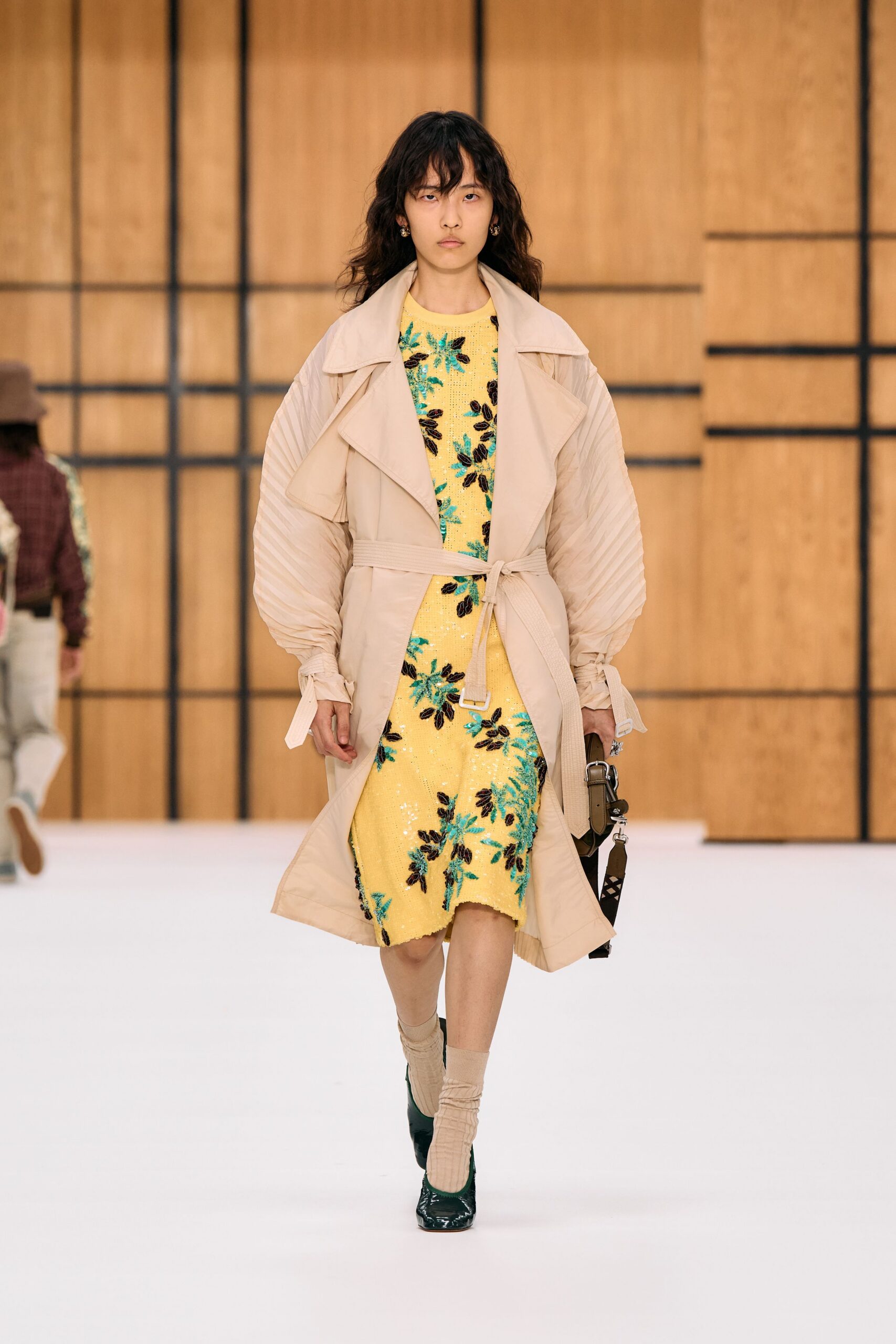 Pale putty trench with accordion-pleated sleeves and a tied webbing belt. Butter-yellow sequined shift, crusted with turquoise and espresso floral beading. Olive shoulder bag with a clipped strap and silver hardware. Beige slouch socks. Dark green patent pumps. Walking into a hotel lobby still carrying garden-party daylight.