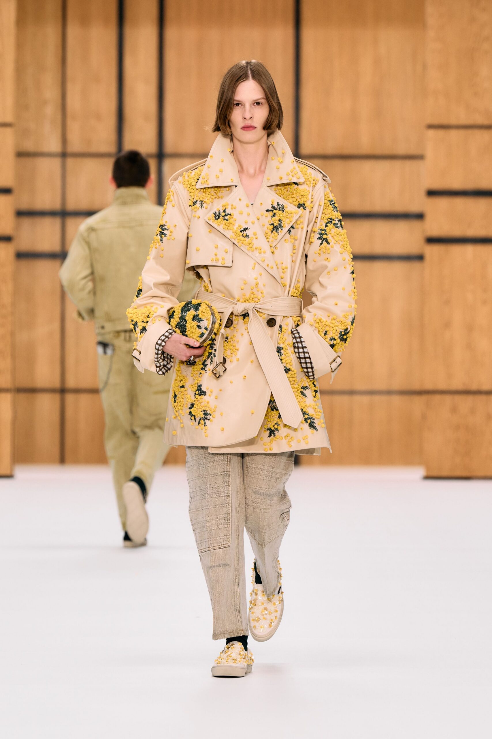 Stone trench coat crusted with yellow mimosa sprigs and beadlike blossoms. Matching oval clutch, densely tufted in the same mimosa clusters. Pale slub-weave trousers with a centered seam and soft break. Cream slip-ons covered in yellow floral tufts. Walking into a conservatory gift shop while pollen still clings to your sleeves.