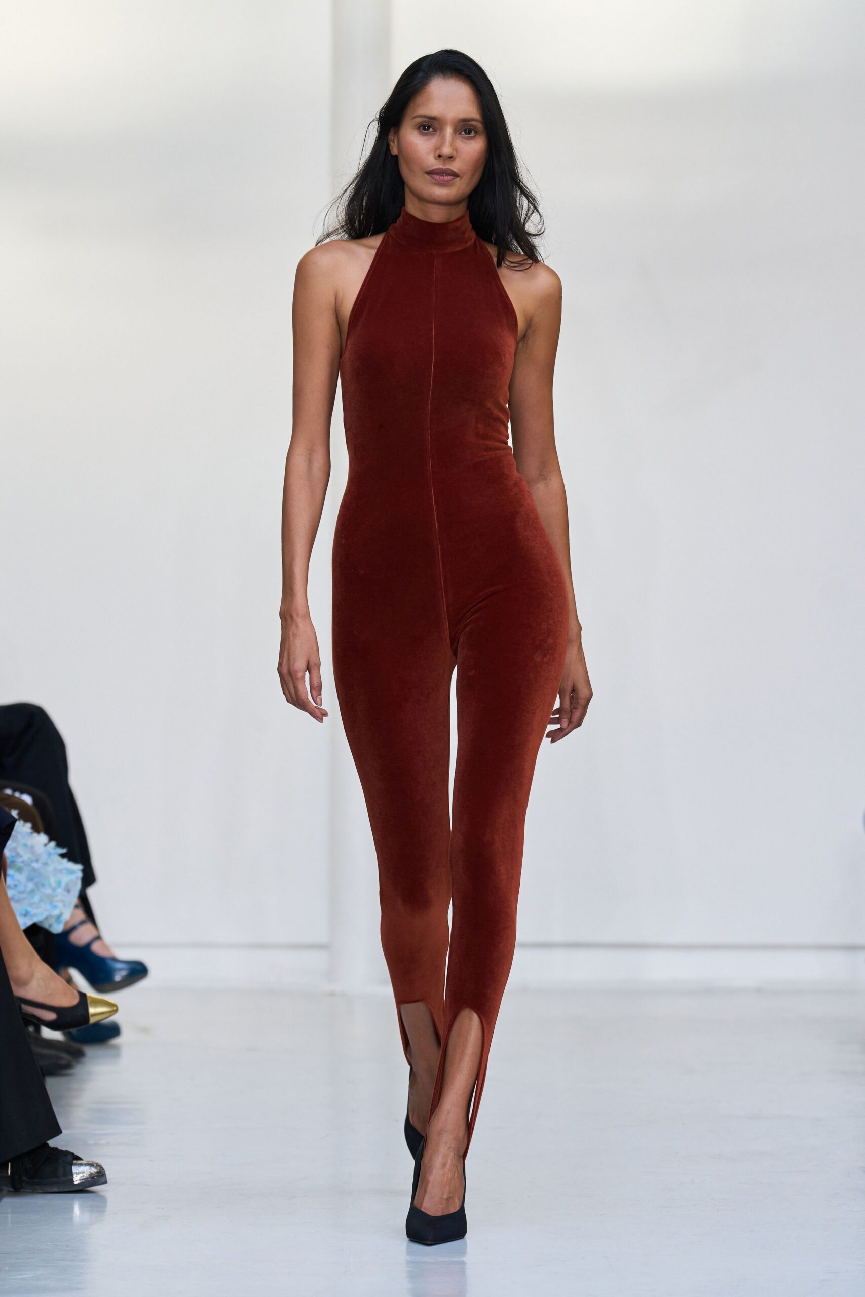 Rust velvet catsuit with a halter mock neck and front seam. Black pointed pumps under split stirrup hems. For crossing a hotel lobby before anyone is fully awake.