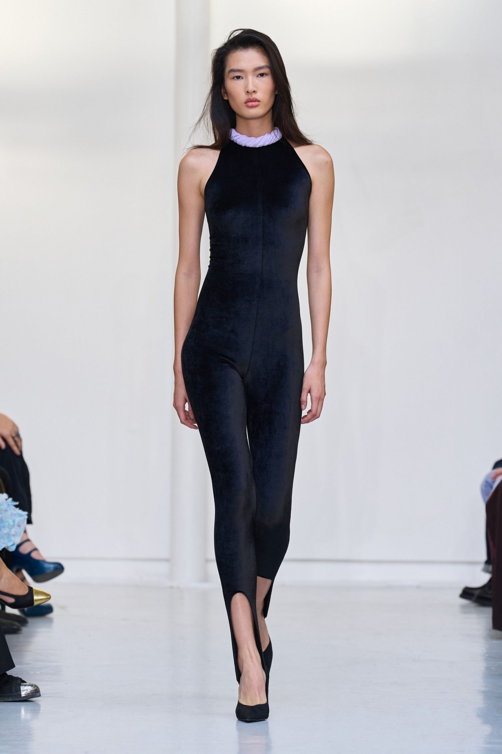 Black velvet halter jumpsuit, center seam running straight to the split hem. Pale lilac twisted collar sitting thick around the throat. Black suede pumps. Museum atrium at noon, your footsteps louder than the fountain.