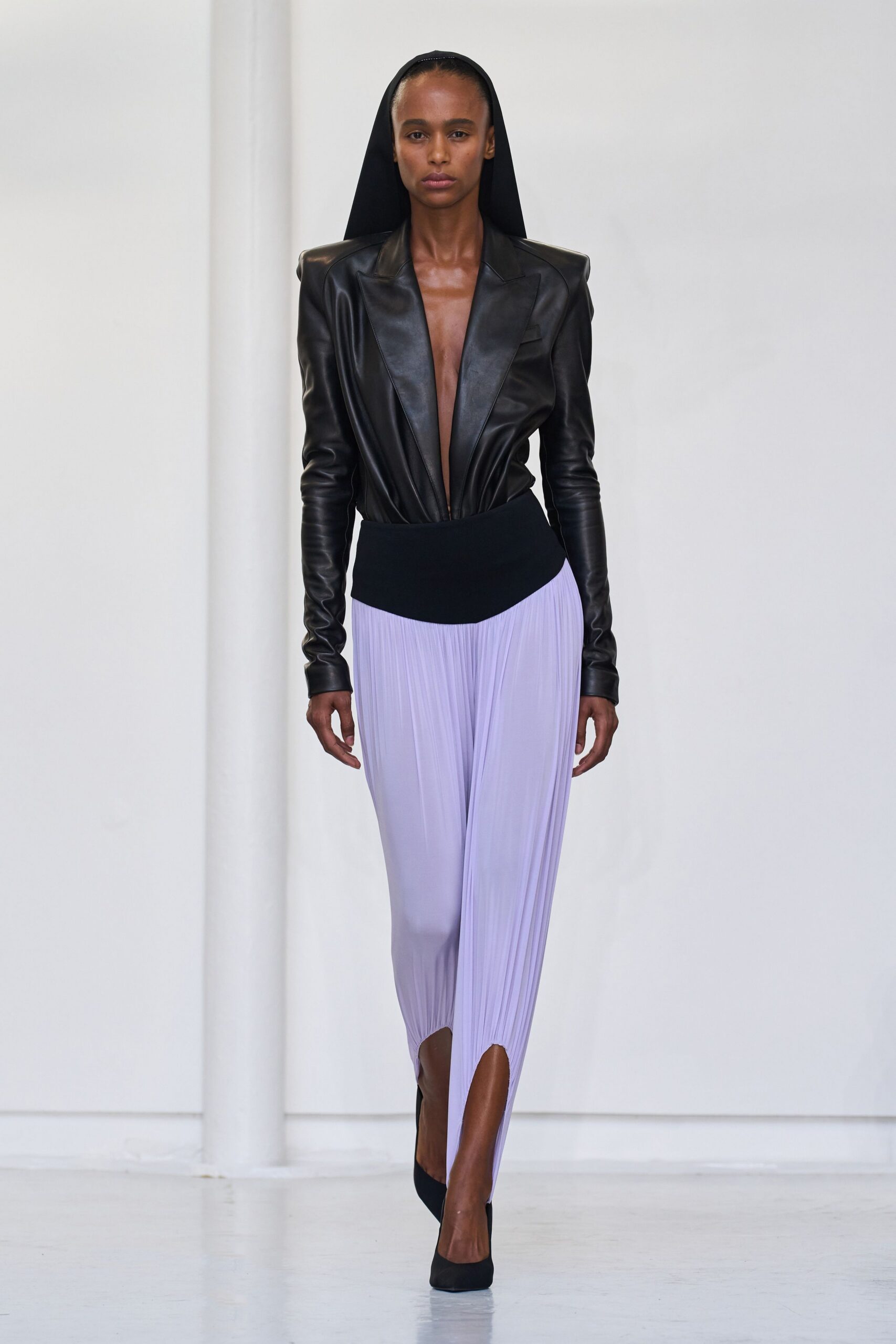 Long black hood falling flat from the crown. Black leather bodysuit with peaked shoulders and a plunging bare front. Lilac micro-pleated trousers under a curved black hip yoke. Black suede pumps. Crossing a silent museum atrium before the doors unlock.