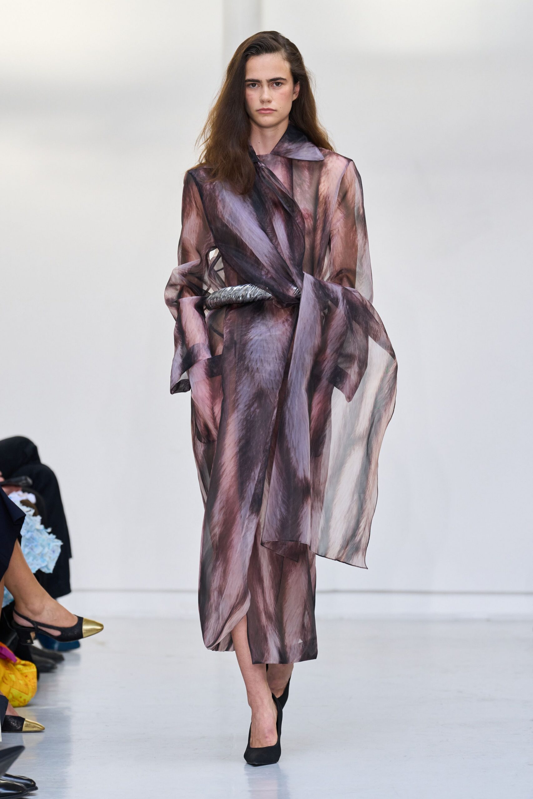 Sheer mauve-and-smoke coatdress, brushstroke print swirling across exaggerated wrap panels. Pewter padded belt knotted low, one end jutting awkwardly outward. Black pumps with a starkly squared toe. Walking into a near-empty museum just after the rain stops.