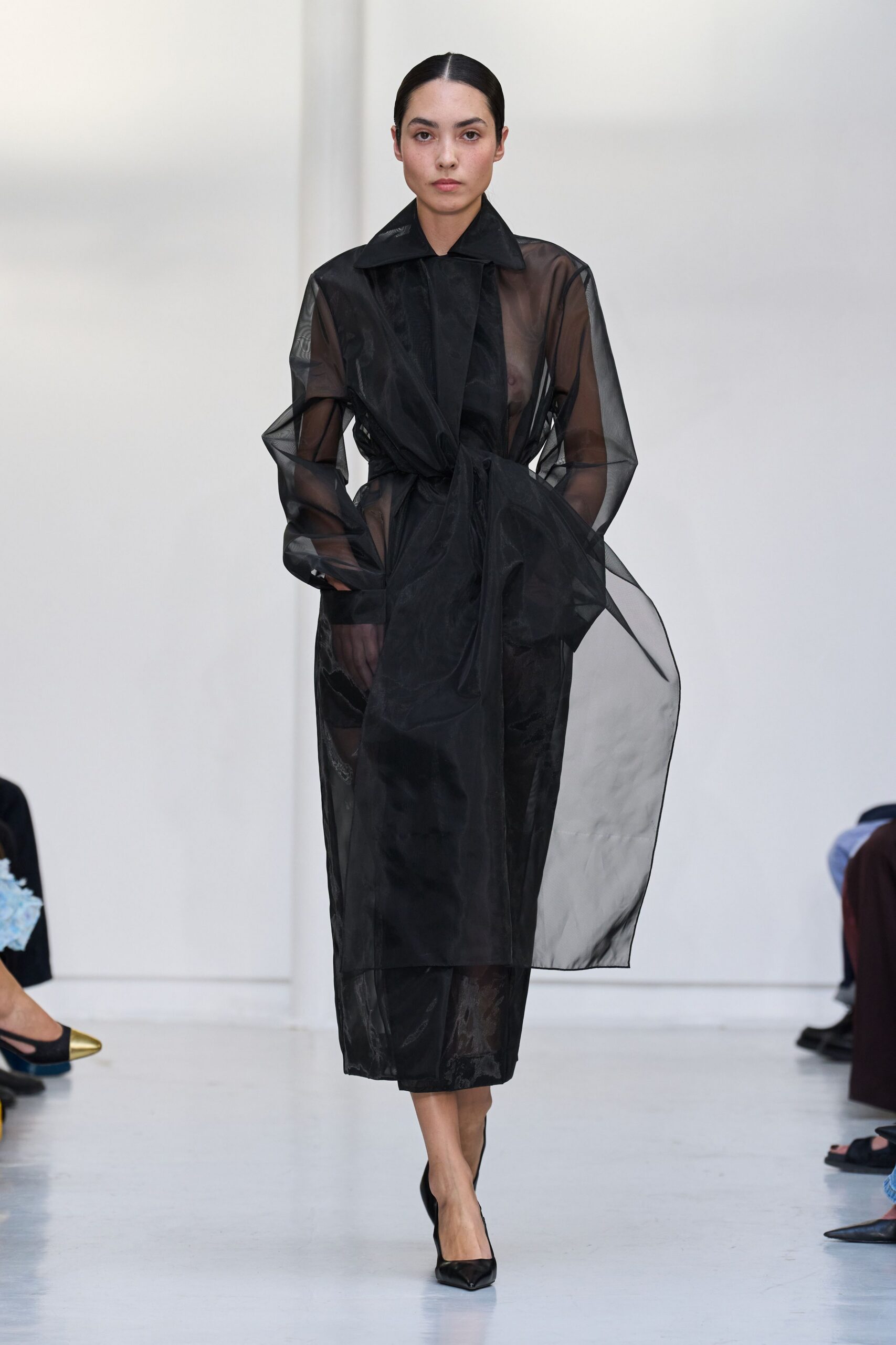 Sheer black organza trench, lapels crisp and one front panel kicked open. Black column dress underneath, its glossy hem ghosting through the organza. Black pointed pumps. For crossing a museum atrium while the rain still sheets off your coat.