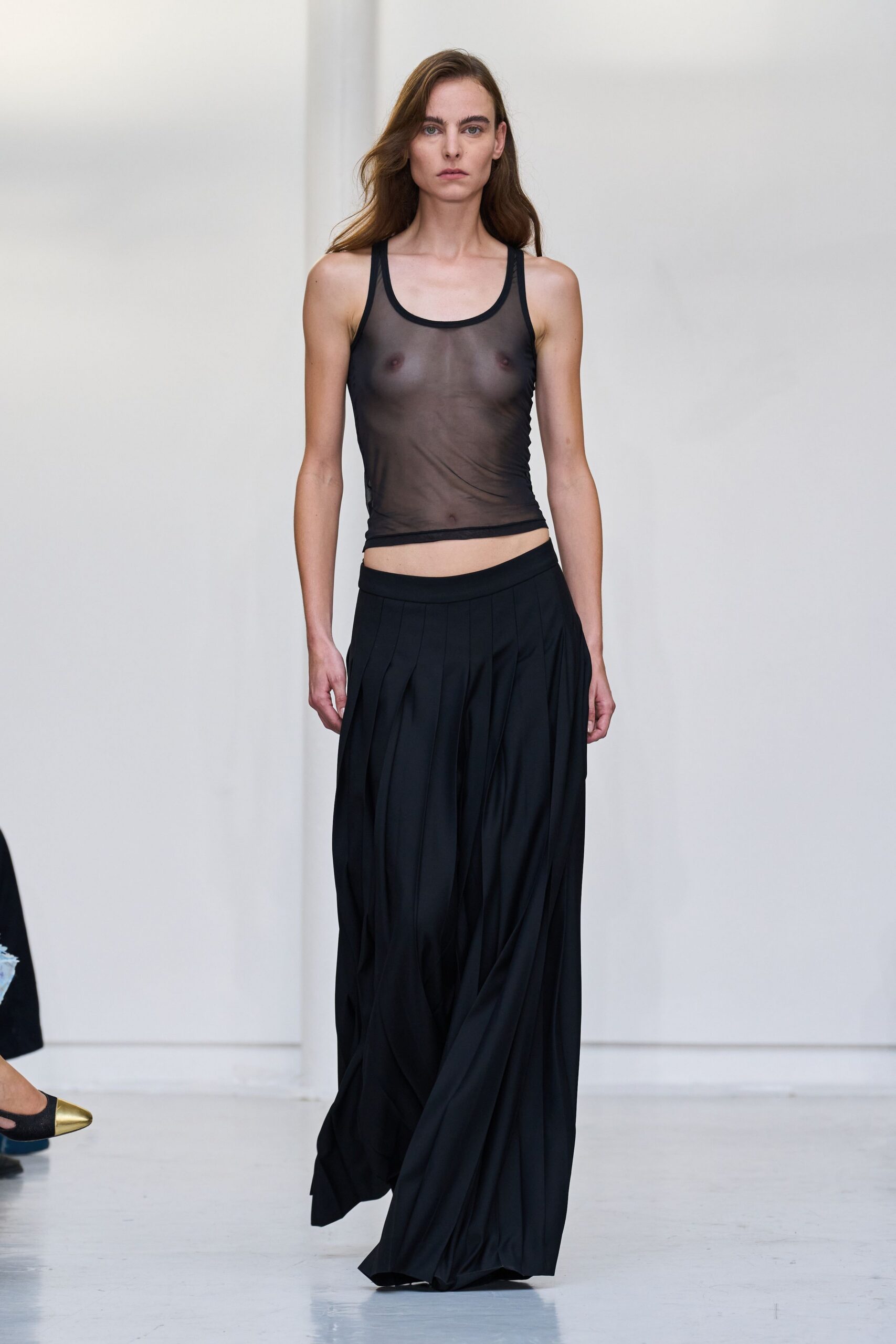 Smoke-black mesh tank, raw-looking hem tipping lower at one hip. Black wide-leg trousers with deep inverted pleats collapsing into soft folds. Walking home barefoot after the coat check lost your dress.