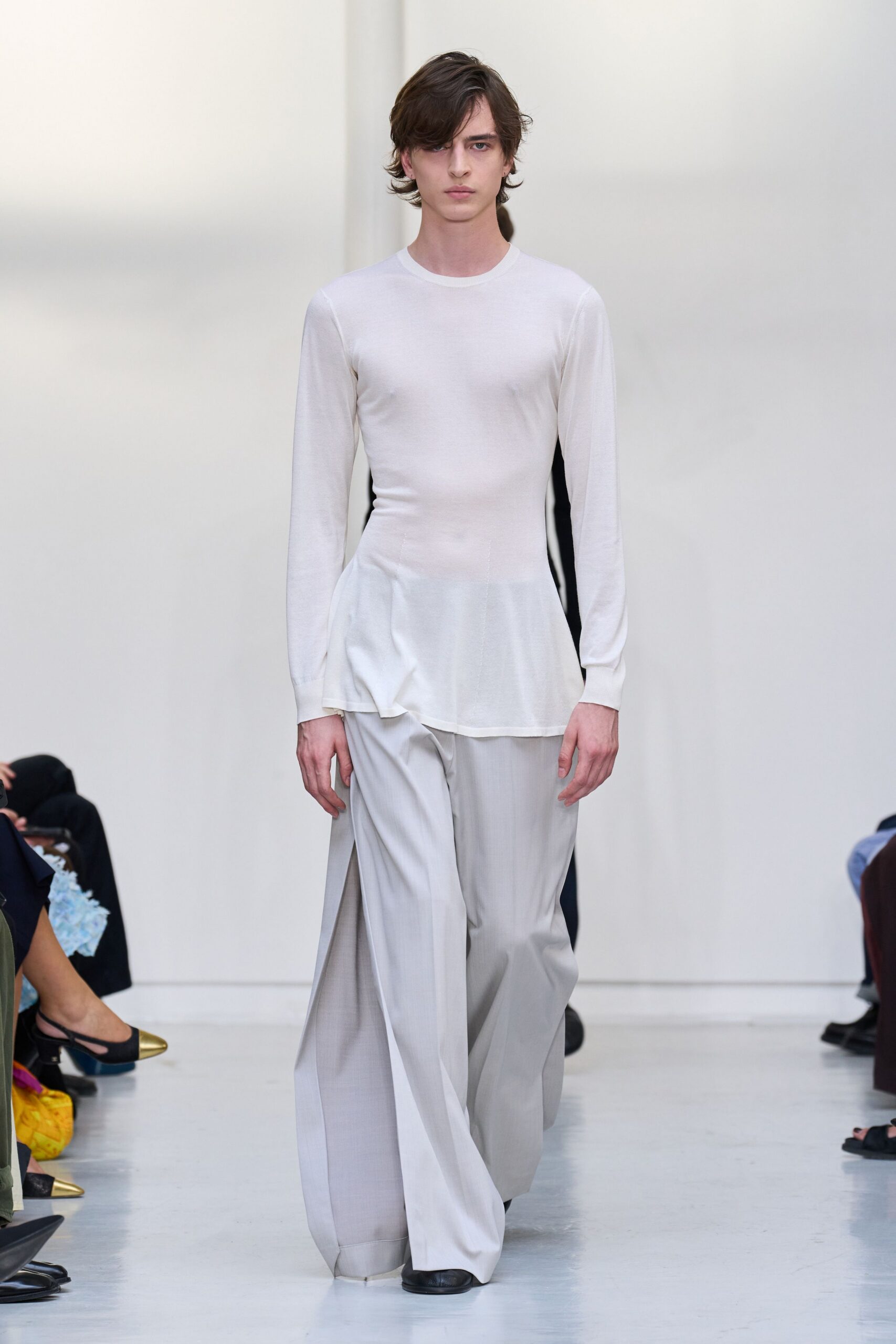Milk-white jersey top, body-skimming with a peplum flare at one hip. Pale dove trousers cut extra wide, one front panel opening into a deep slit. Black square-toe shoes disappearing under the pooled hem. Crossing a museum atrium before the air-conditioning fully wakes up.