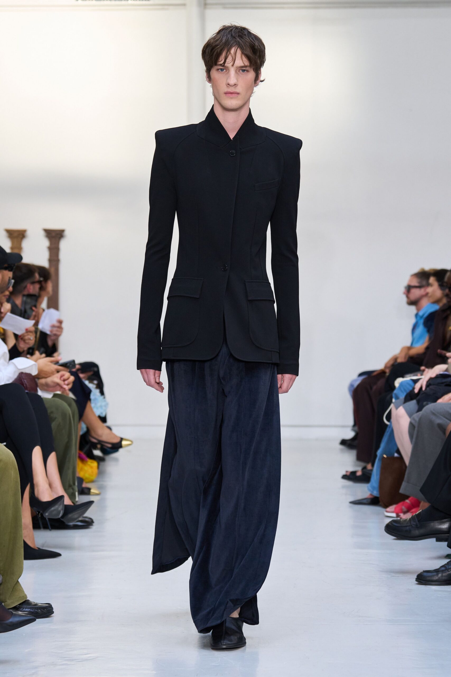 Black tailored jacket with sharp pagoda shoulders and curved welted pocket flaps. Inky fluid trousers falling from a dropped crotch into a pooled hem. Black square-toe shoes, vamp barely peeking under the break. Crossing a museum atrium before the security gates unlock.