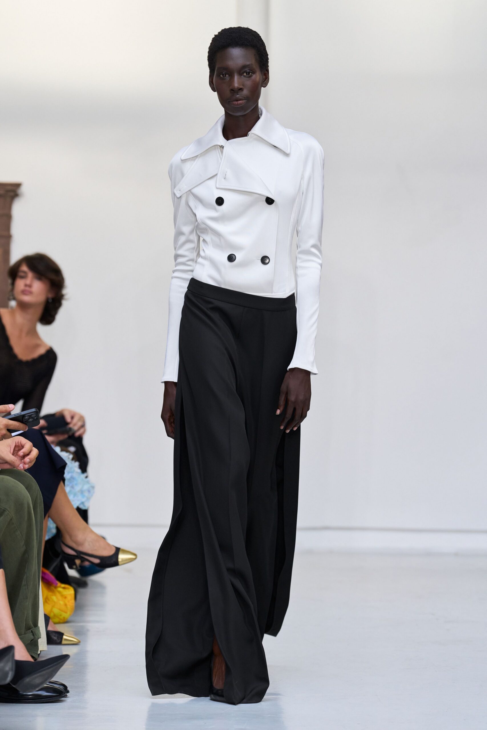 Chalk double-breasted jacket with a skewed storm flap and black buttons. Black wide-leg trousers falling in long creases over the foot. Black pointed heels barely visible under the hem. For crossing a marble lobby before the offices unlock.