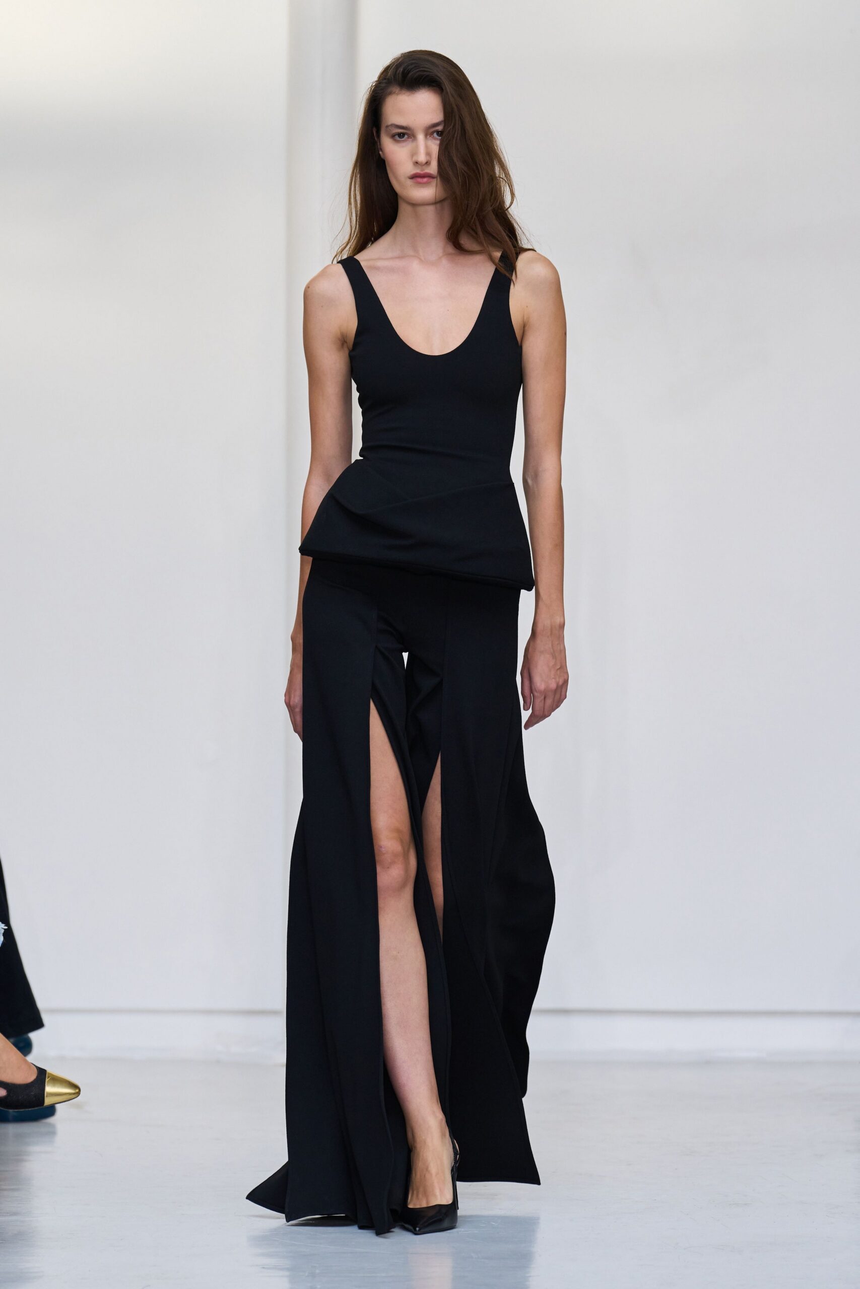 Black stretch-knit tank, scooped low and cut with a stiff peplum flare. Floor-length black skirt split to the thigh, panels falling open with each step. Black satin pointed pumps. For crossing a silent lobby after midnight with the keycard already in hand.