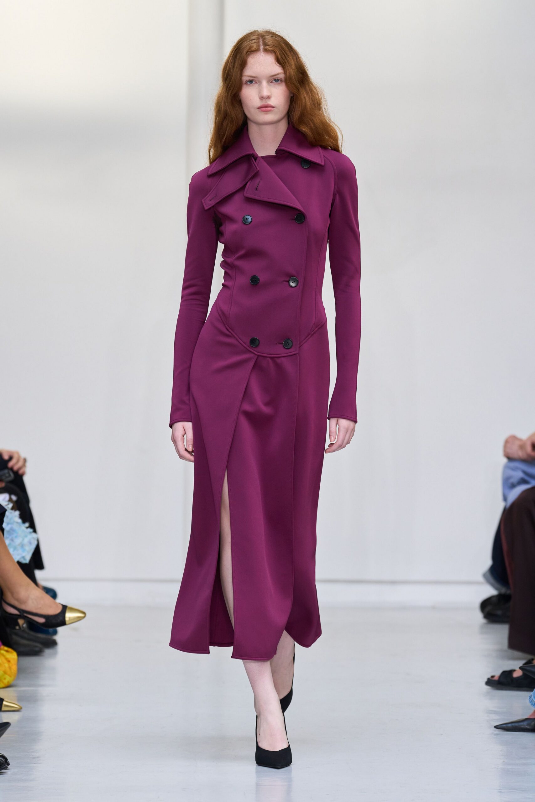 Magenta double-breasted trench dress in dense jersey, one lapel tipped askew. Black pumps with a bare, almost hidden vamp. For crossing a hotel lobby before anyone has ordered breakfast.