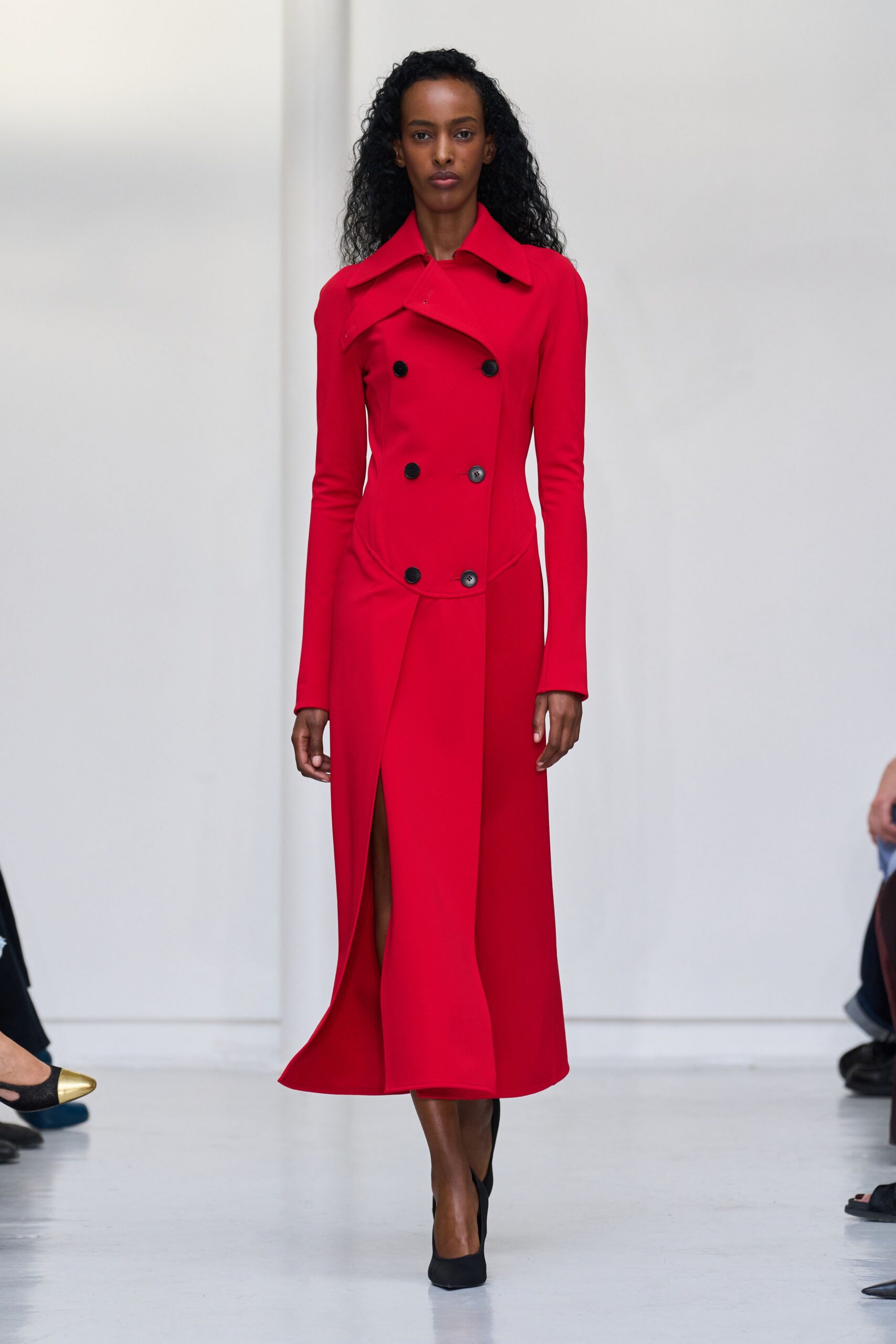 Scarlet double-breasted coat dress, curved welt seam and wide undercollar. Black pumps, squared toes peeking from the front slit. For entering a courthouse after the rain has finally stopped.
