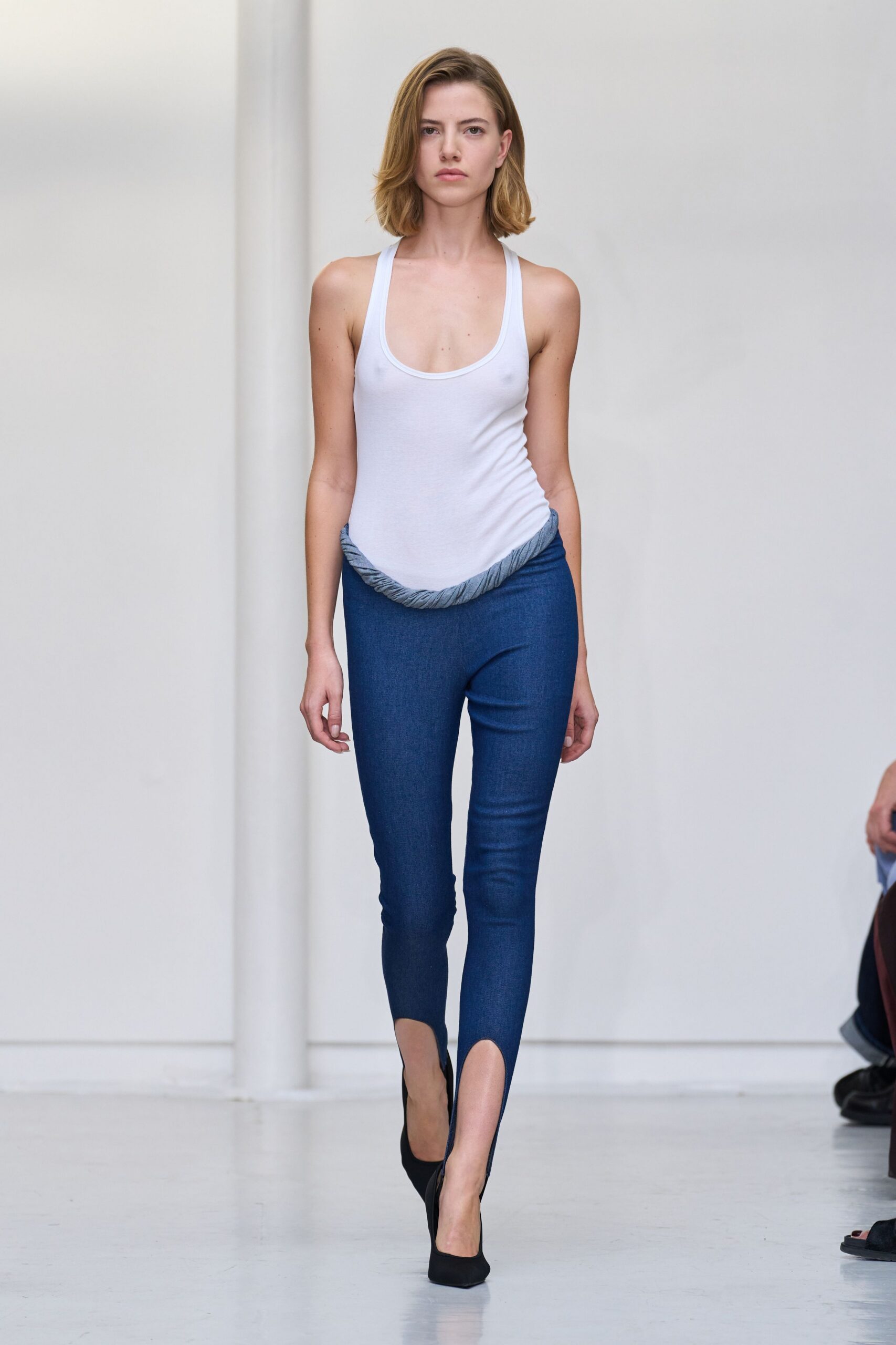 White stretch bodysuit with a scooped racerback neckline. Dark indigo leggings with a twisted grey waistband and split stirrup hems. Black pumps in matte suede. For crossing a spotless loft before anyone has had coffee.