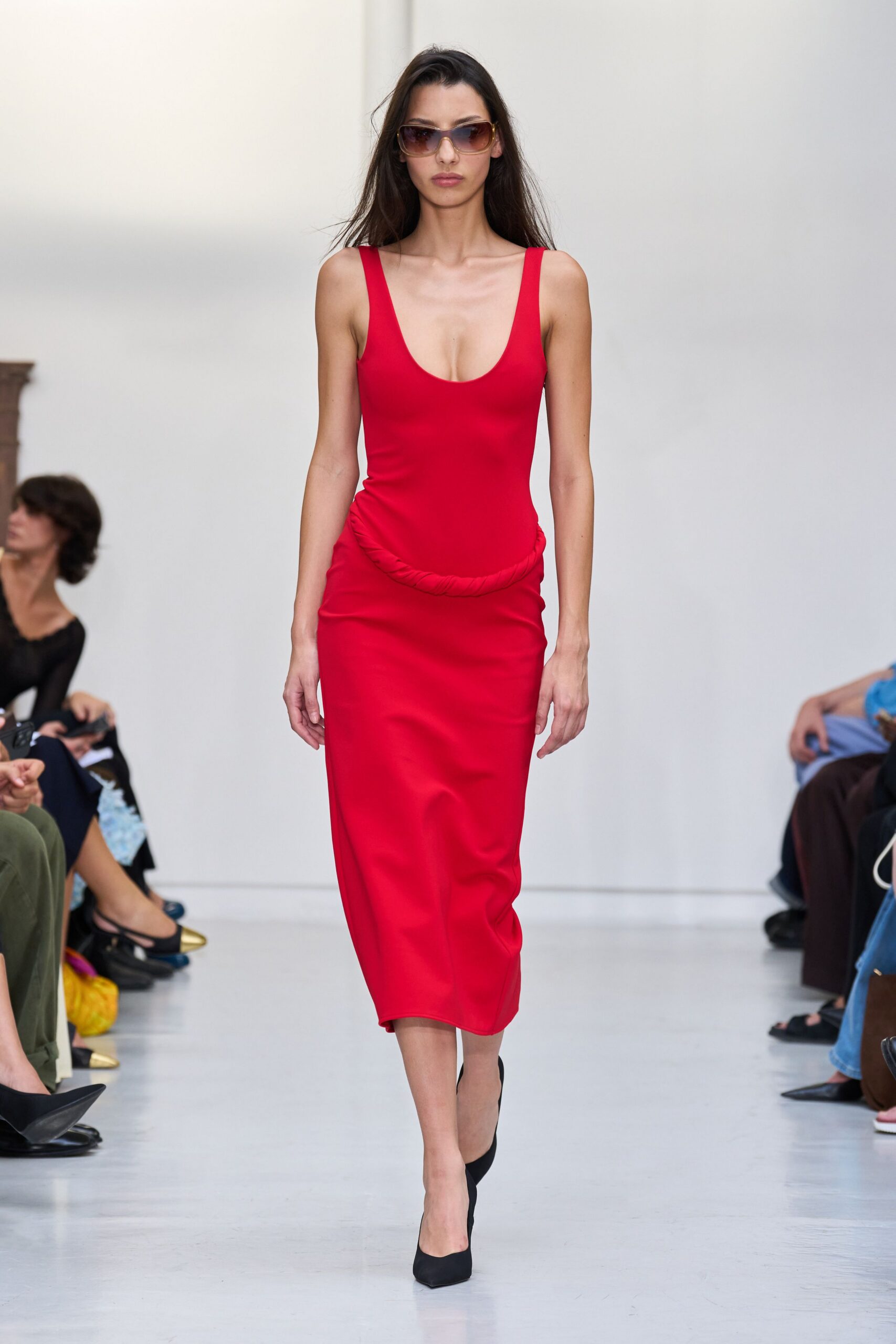 Amber-gradient wraparound sunglasses with a thick translucent frame. Scarlet column dress in dense stretch jersey, a twisted rope seam slung low at the hips. Black pointed pumps in matte suede. For walking into a hotel lobby before anyone knows why you're there.