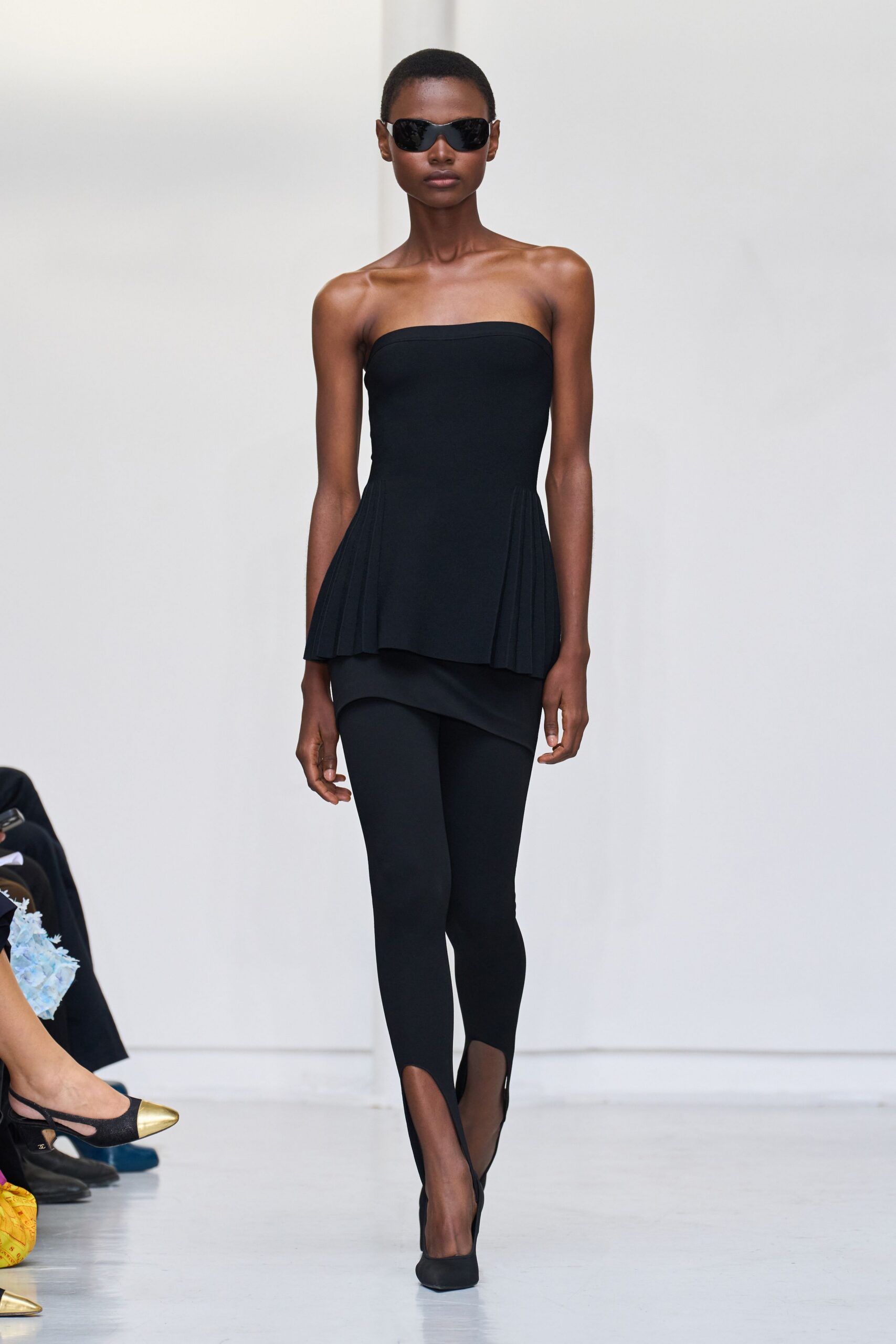 Wraparound black lenses with a narrow glossy frame. Strapless black top, side pleats opening from the hipline. Short black underskirt cut on a sharp diagonal. Opaque black stirrup leggings hugging into the arch. Black pumps with a softly squared toe. For crossing a white lobby before anyone else is fully awake.