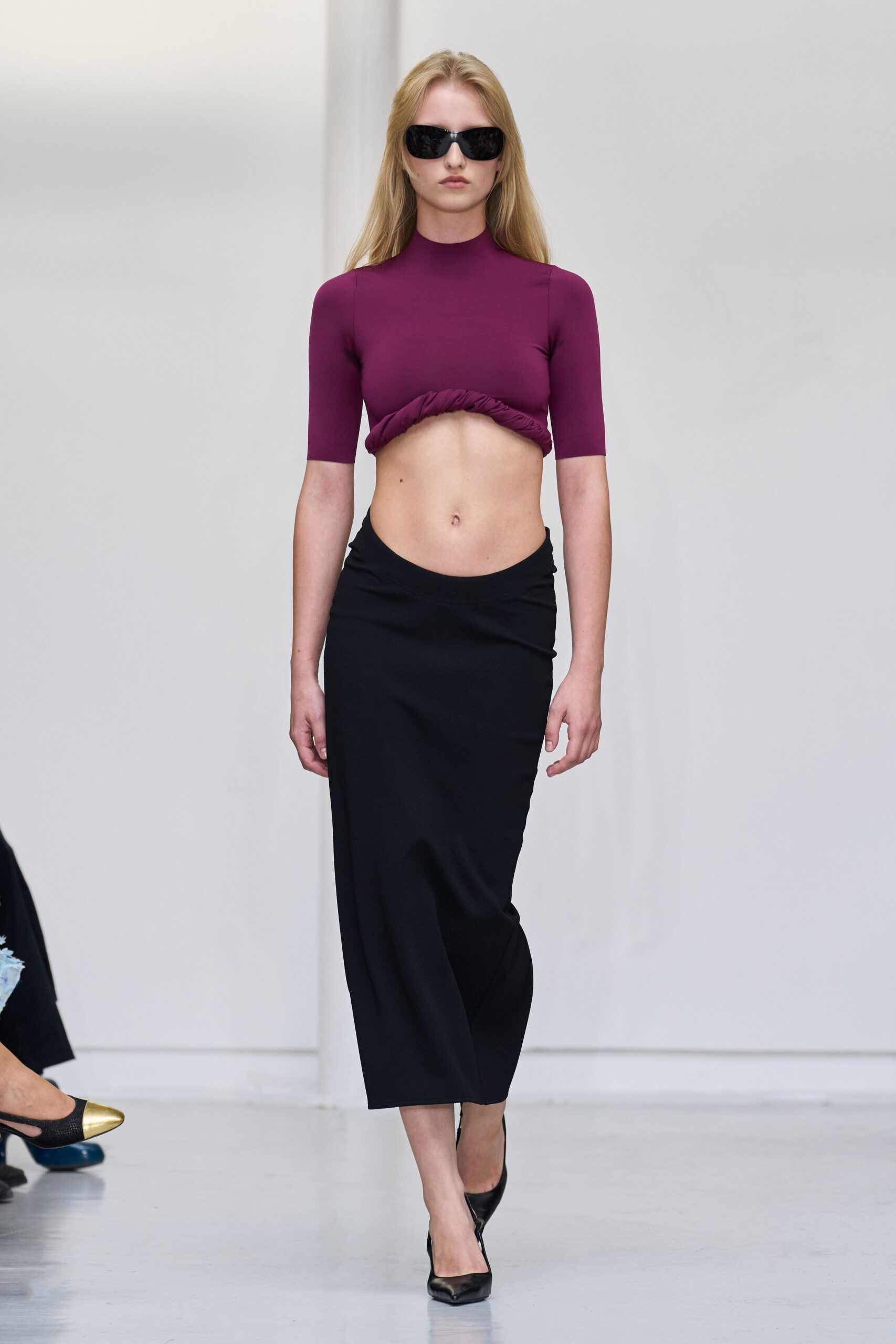 Glossy black wraparound sunglasses. Plum stretch jersey crop top, ruched underbust hem curled upward. Black column skirt with a dipped waistband and slight twist at hip. Black pointed pumps. Walking from the hotel lobby to breakfast after sleeping in yesterday's makeup.