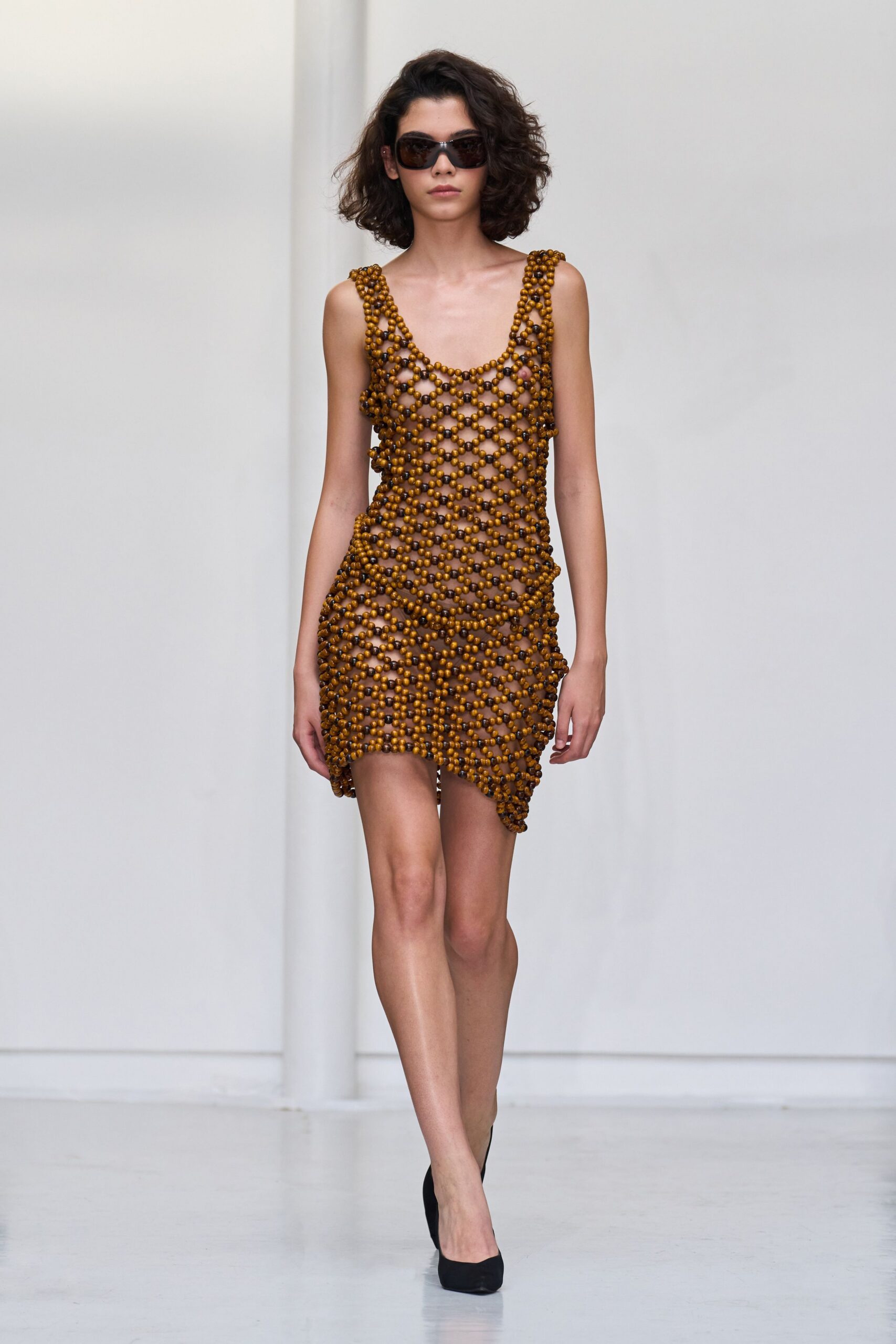 Dark wraparound lenses in a broad brown frame. Open lattice mini strung with honey-brown wooden beads, hem dipping uneven at one thigh. Black pumps with a plain matte finish. For crossing a hotel lobby still damp from the pool.
