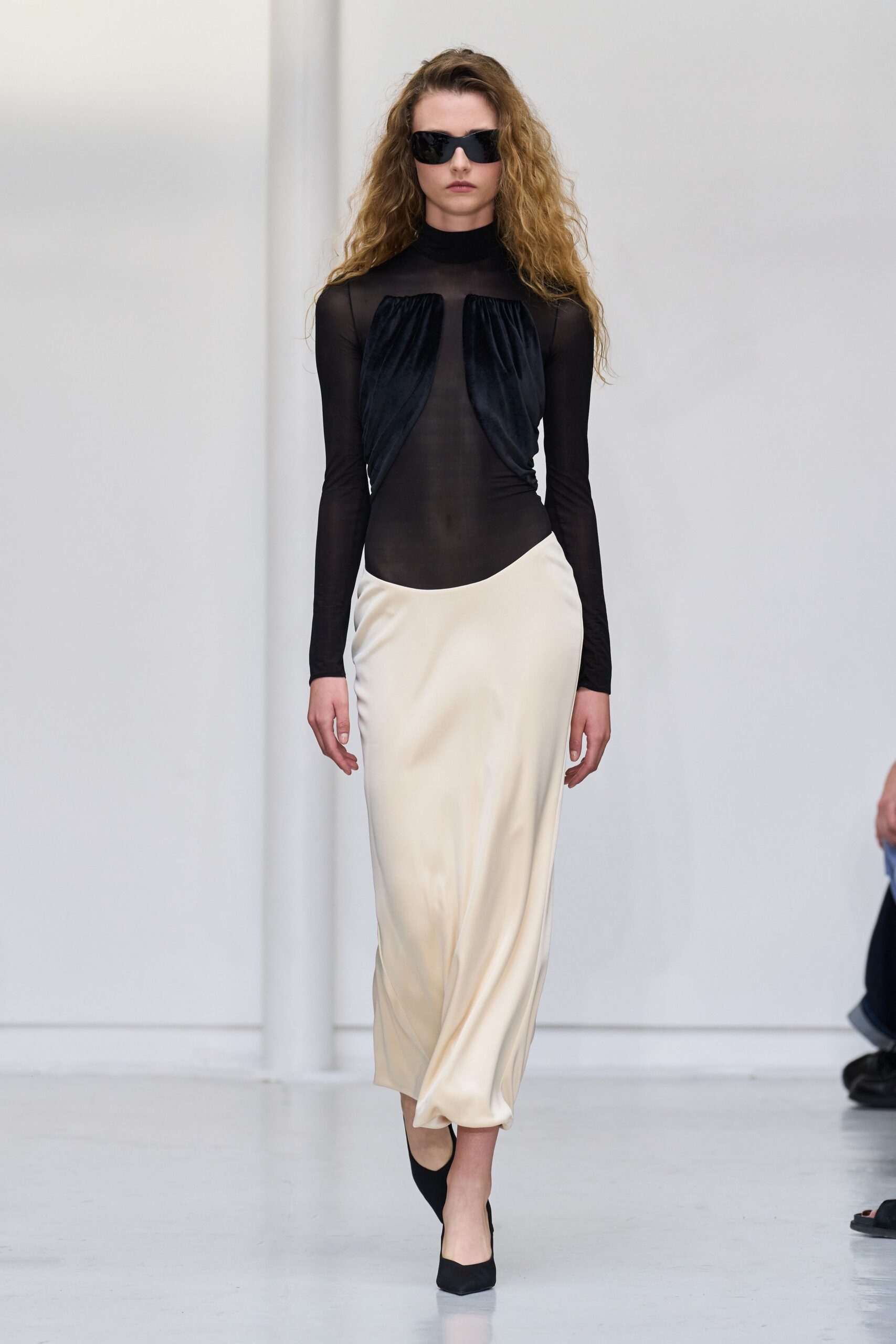 Wraparound black sunglasses with a sharp, visor-like curve. Sheer black mesh bodysuit, velvet panels gathered from the high neck. Ivory satin column skirt with a low curved waist and pooled hem. Black pointed pumps. Walking through a white atrium after changing out of stage clothes.