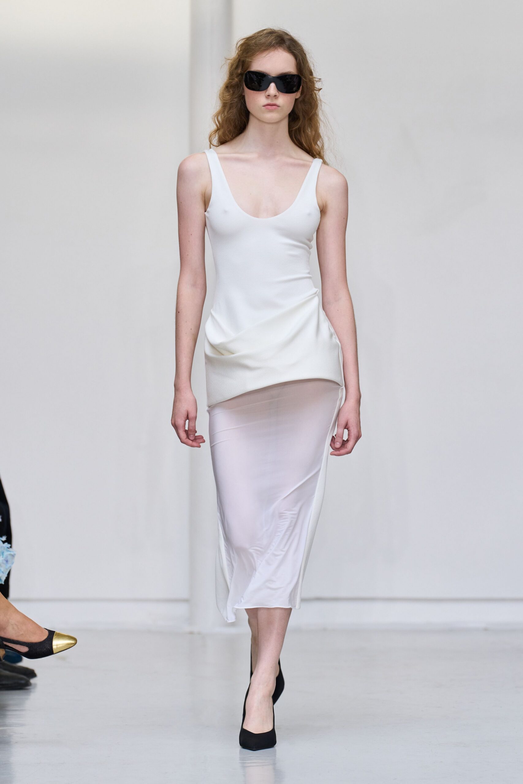 Oversized black wraparound sunglasses. Ivory stretch slip dress with a scooped neck and sheer lower panel. Black pumps with squared vamps. Walking the long white corridor after changing out of swimwear.