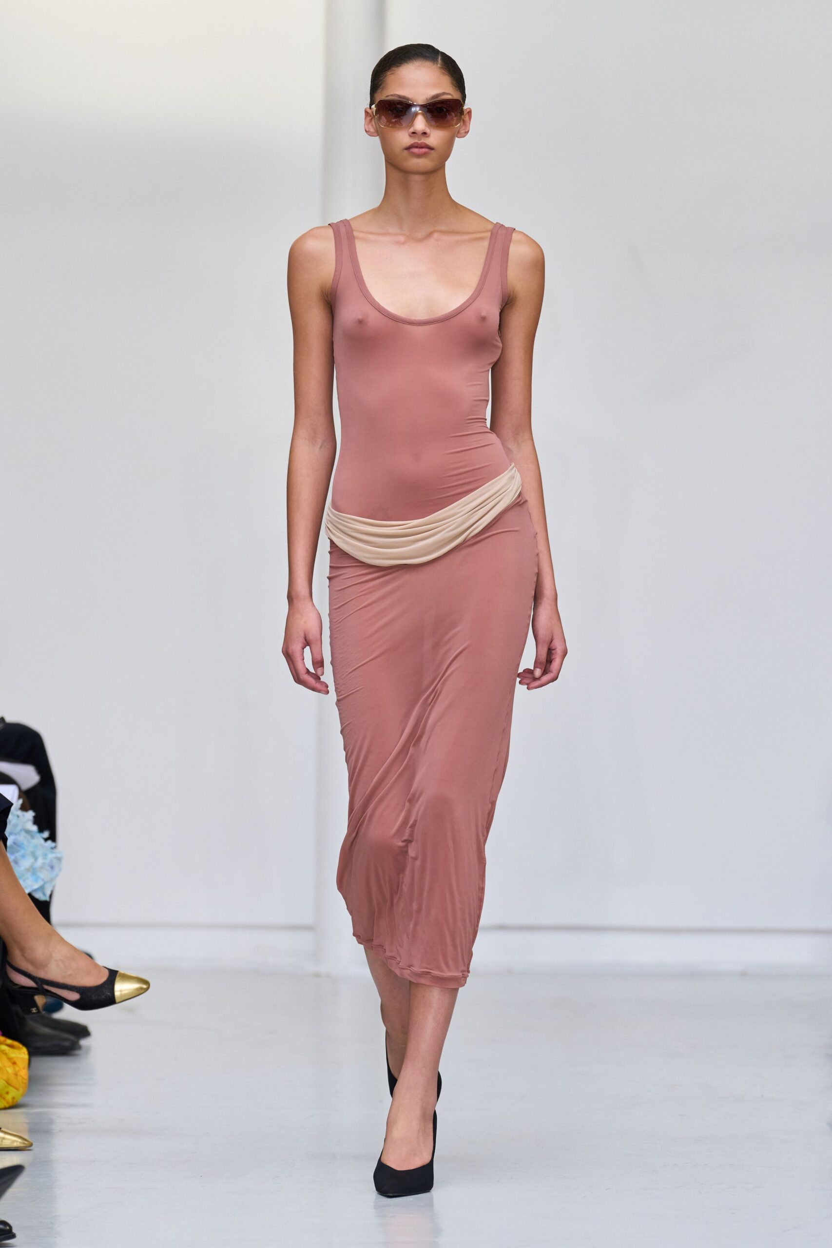 Oversized brown gradient shield sunglasses. Dusty rose jersey tank dress, clingy and slightly sheer through the torso. Cream jersey panel slung low, softly ruched across one hip. Black pumps with a plain closed toe. Walking back from the hotel pool after the towel's already damp.