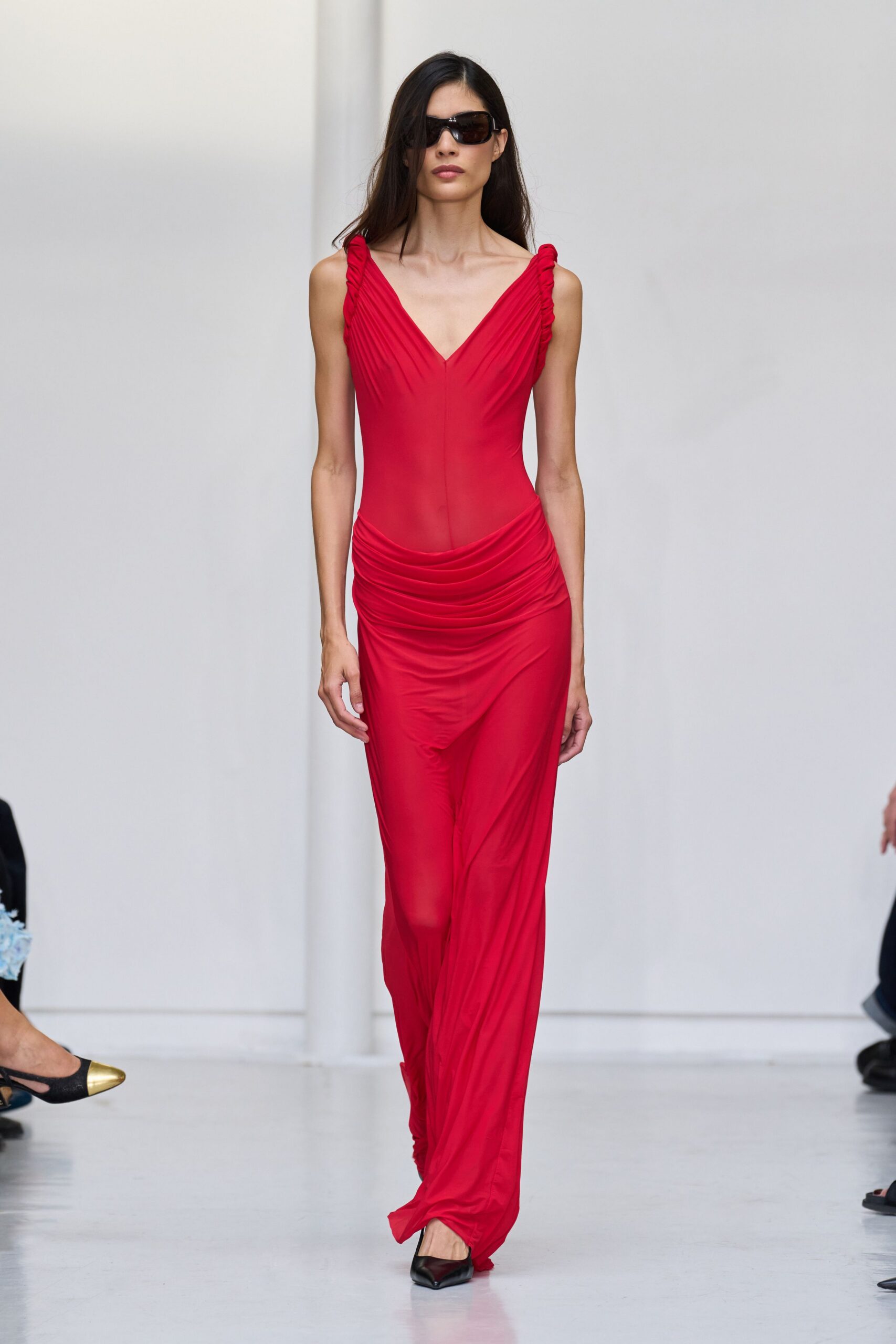 Oversized black wraparound sunglasses with a thick glossy temple. Poppy-red jersey column dress, ruched shoulders and a softly dropped waist. Black pointed pumps barely visible beneath the dragging hem. Whatever you wear to collect keys from an ex in daylight.