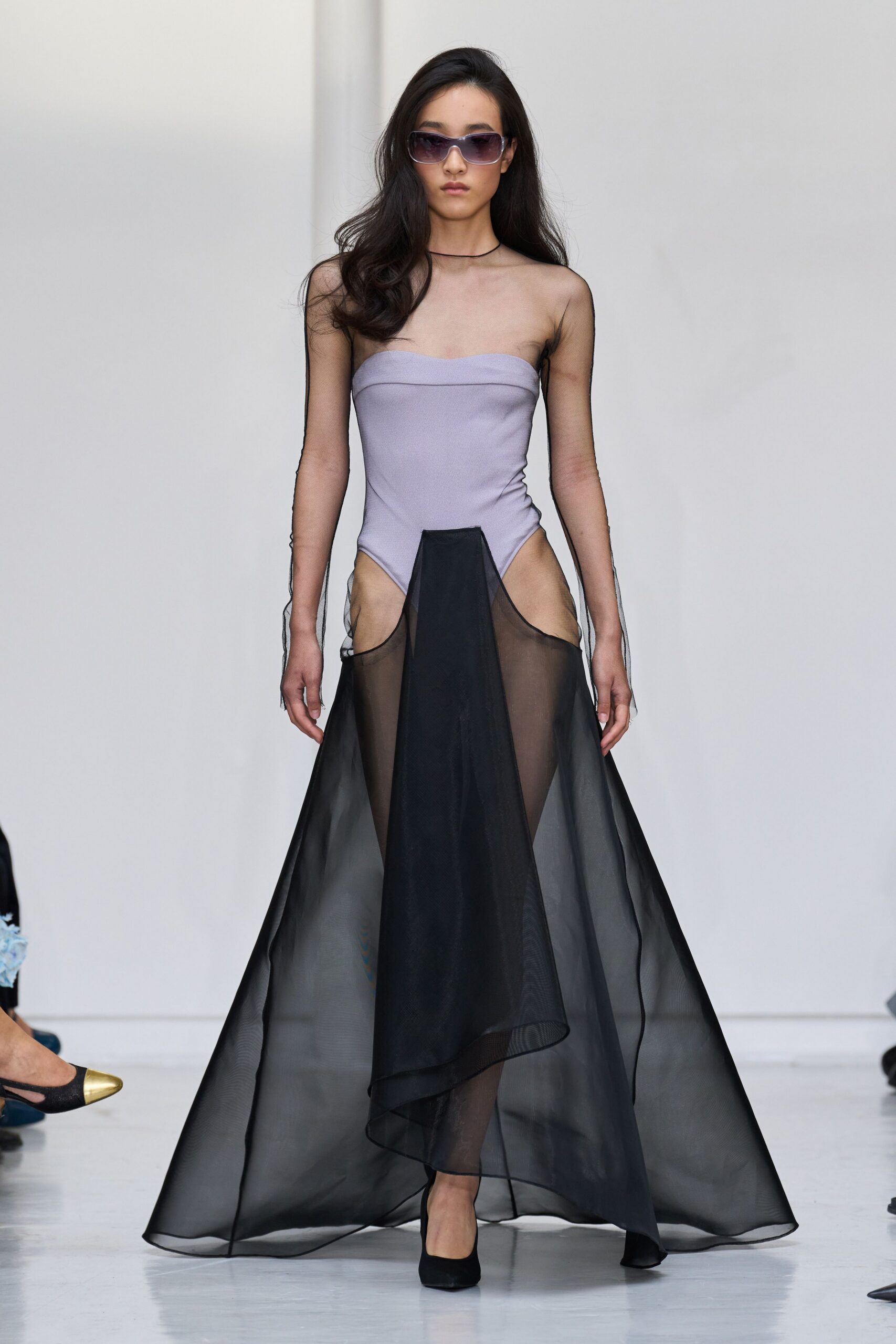 Tinted lilac shield sunglasses with a softly squared frame. Thin black cord sitting close against the collarbone. Dove strapless bodice with sheer black mesh sleeves and a floating organza overskirt. Black suede pumps, plain and sharply pointed. Crossing a museum atrium before the fountains switch on.