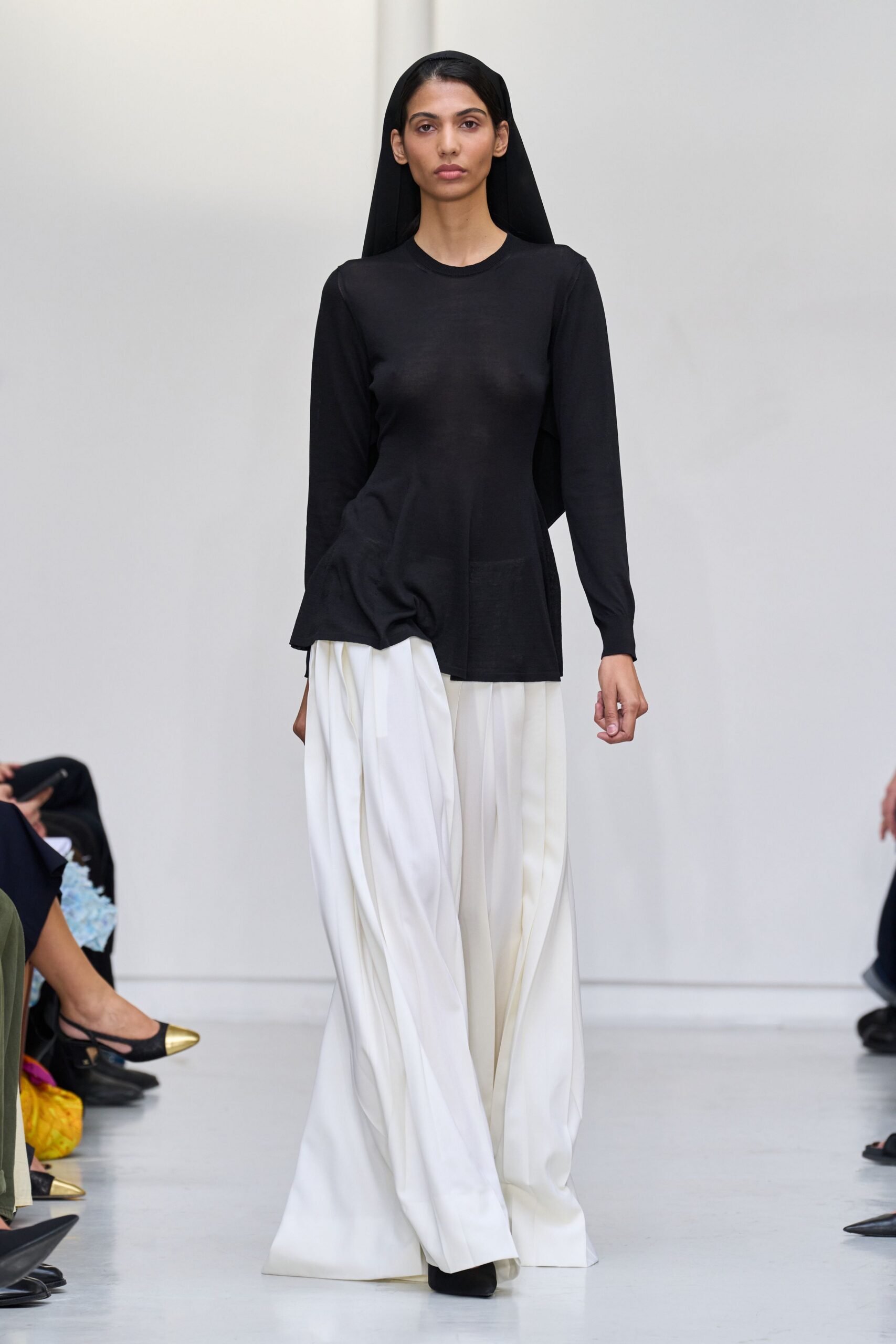 Black jersey veil falling straight past the shoulders. Fine black knit top, one side draped into a low twisted peplum. Ivory wide-leg trousers in fluid crepe, deep folds breaking over the shoes. Black pointed boots, mostly hidden under the pooling hem. Crossing a museum atrium while the marble floors are still being mopped.
