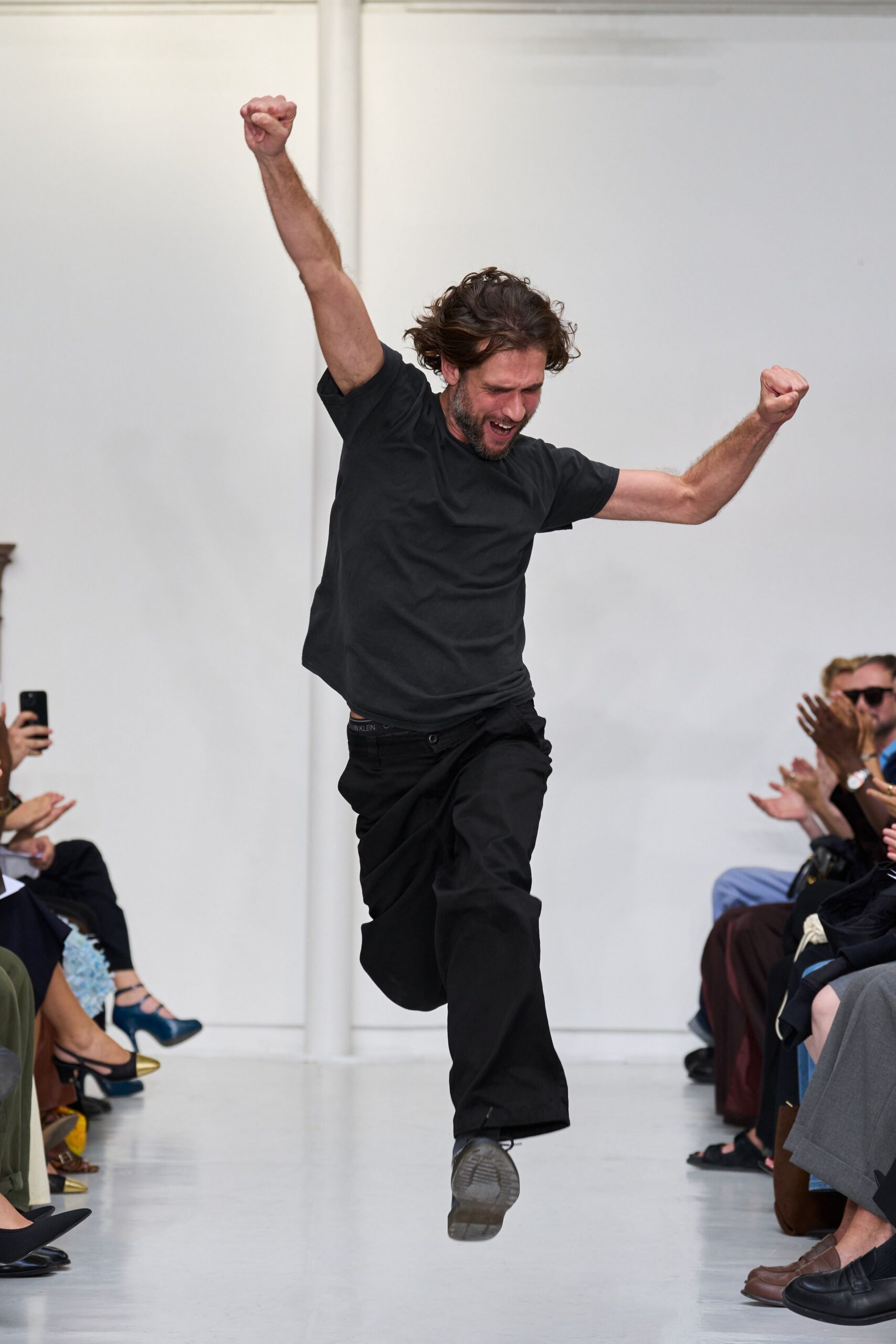 Washed black cotton tee with a soft, slightly collapsing crewneck. Loose black trousers, plain front, waistband dipping to show logo elastic. Black lace-up shoes with greyed, worn soles. Running down the hallway after hearing your name called at the end.
