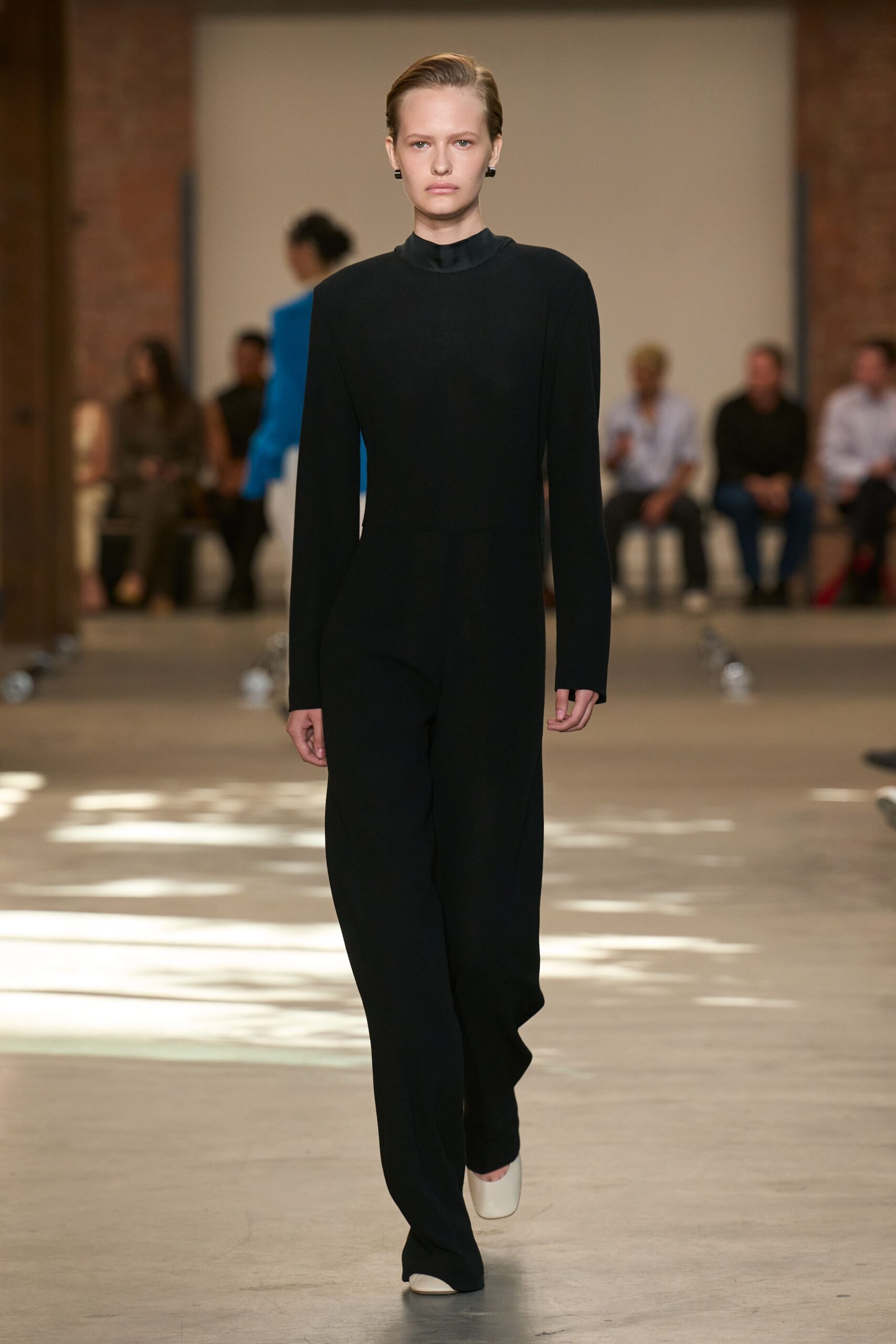 Black crepe jumpsuit with a satin stand collar and long narrow sleeves. Small black rectangular studs. Milk-white square-toe shoes peeking from the pooling hem. Crossing a museum atrium before the first school group gets loud.