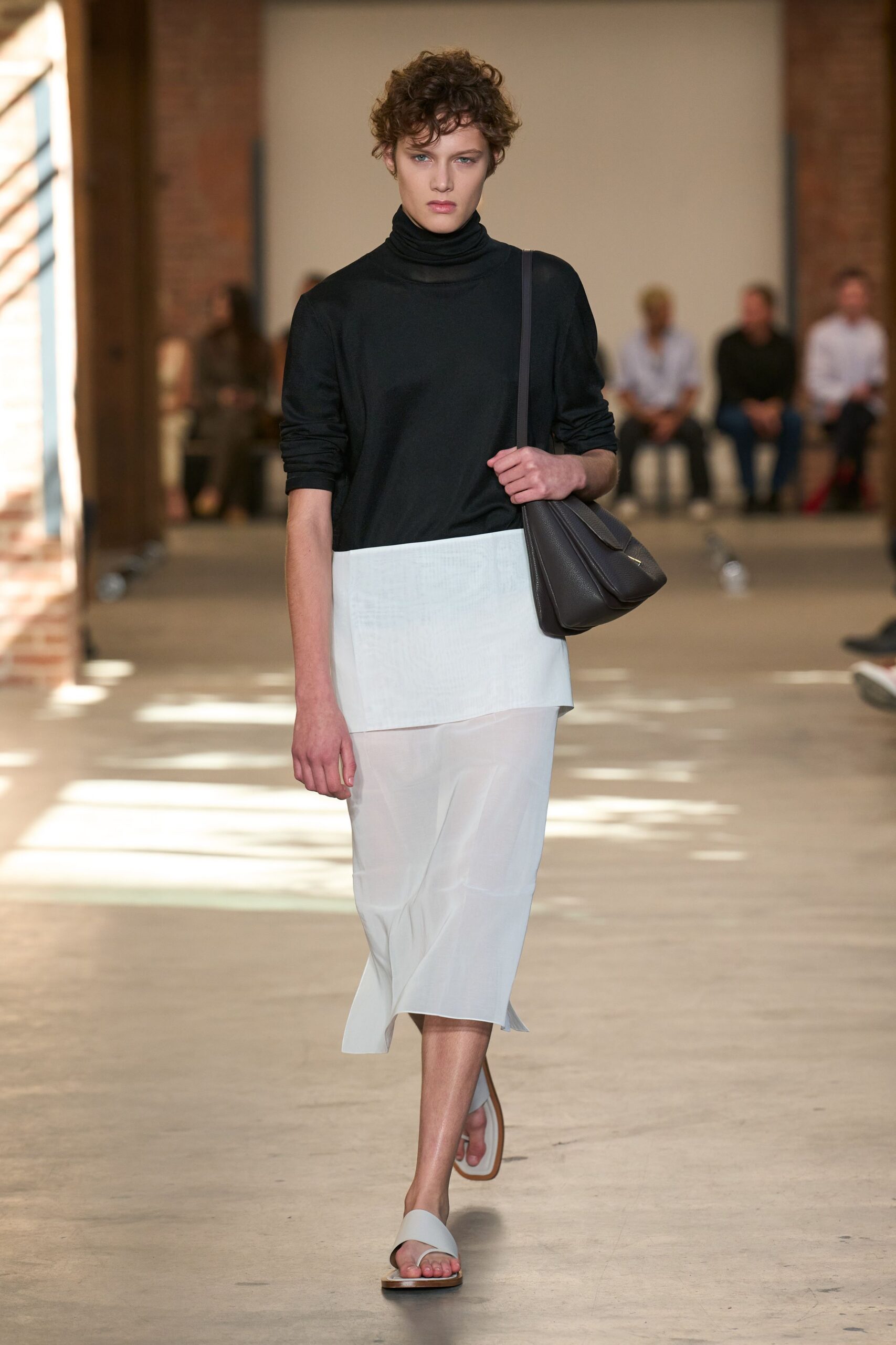 Black fine-gauge turtleneck with a doubled, slouching neck. White wrap skirt layered over a sheer calf-length underlayer. Dark taupe pebbled leather, folded in two soft gussets. Pale thong sandals with a squared wooden sole. Crossing a museum atrium before the café queues start.