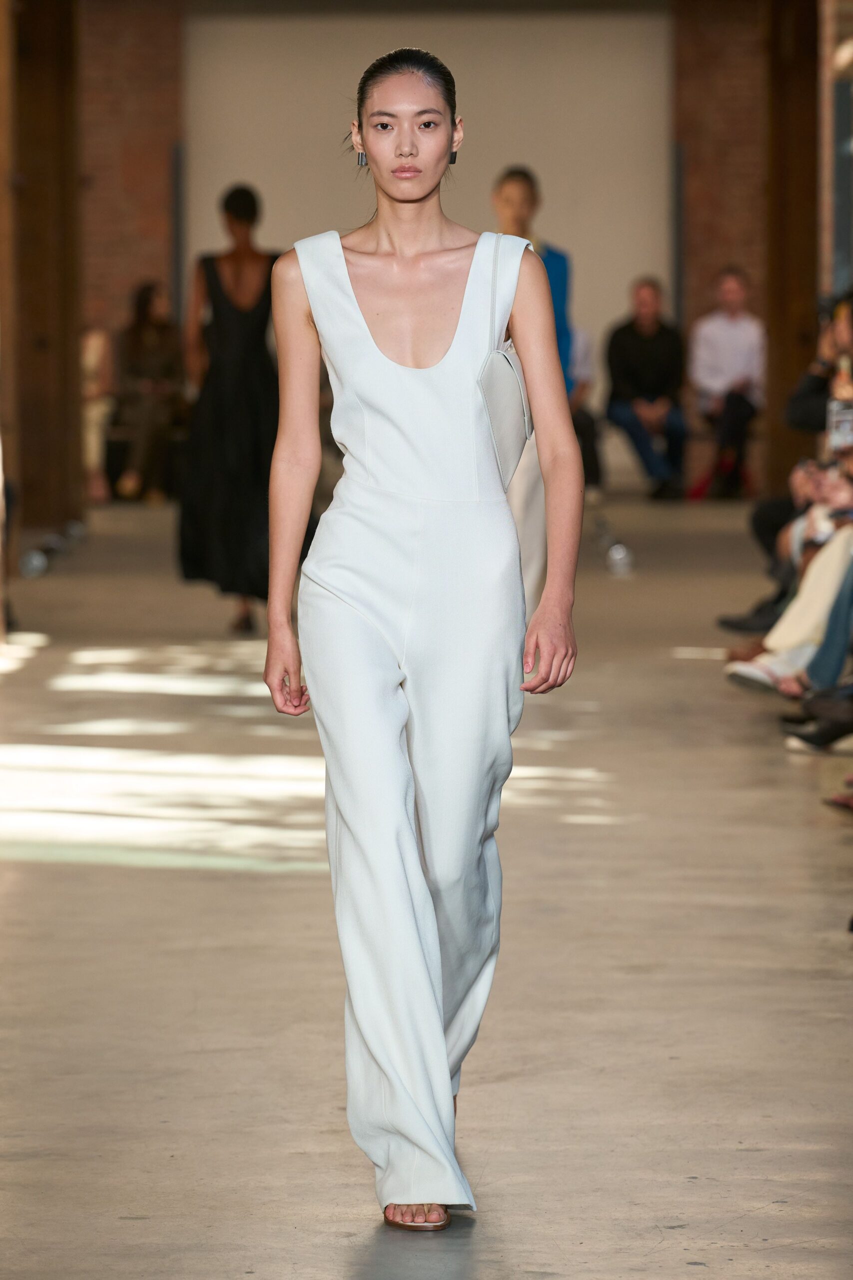 Powder-blue crepe jumpsuit with a scooped neckline and pressed center seams. Matching pale leather shoulder bag tucked tight under the arm. Barely there flat sandals, square sole peeking past the hem. Short black rectangular drops. Crossing a museum atrium just after the air-conditioning kicks in.
