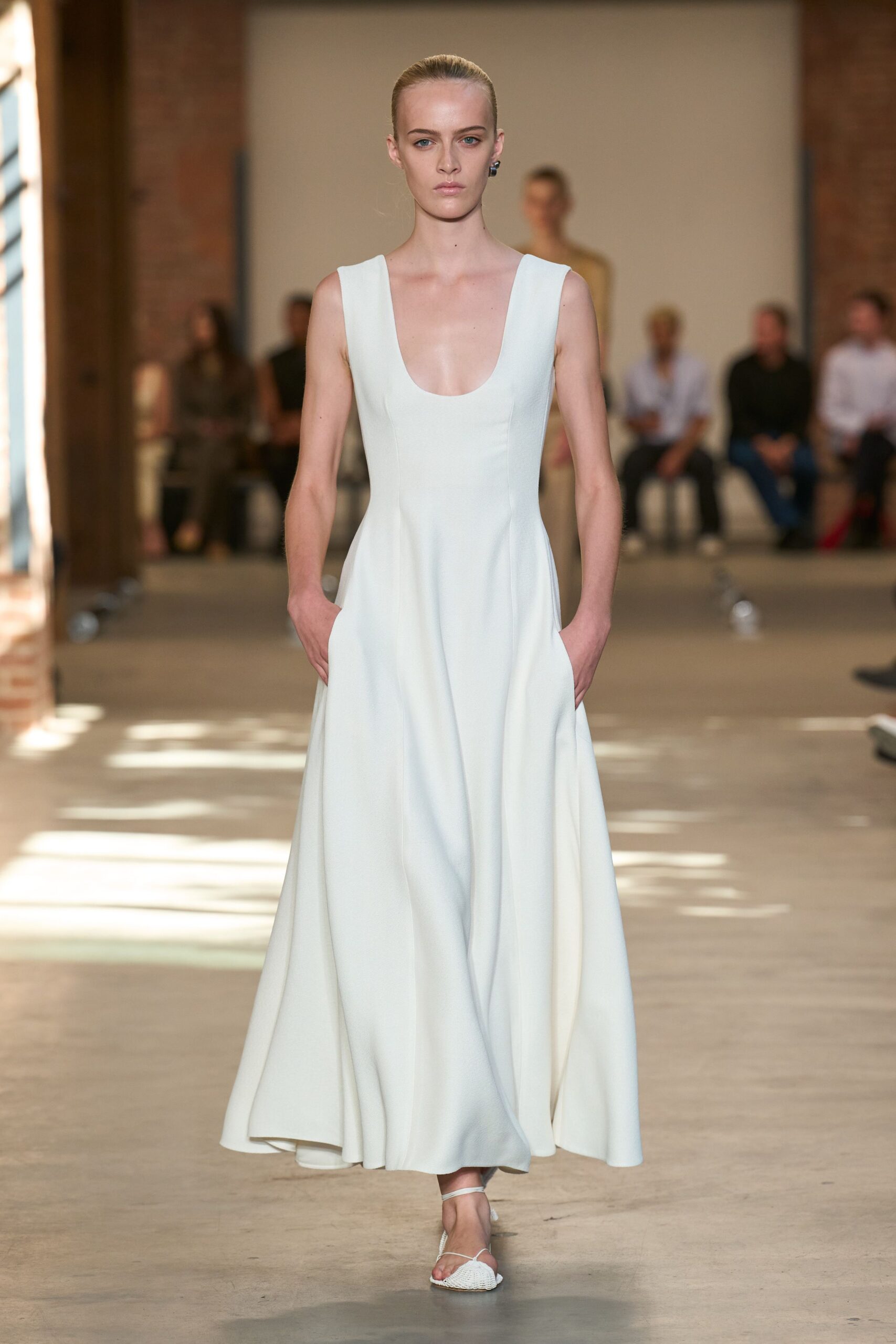 Small black stud earrings, glossy against the slicked-back hair. Ice-blue floor-length dress in dense crepe, scooped low and seamed through the waist. White thong sandals with a woven square toe panel. Sun pooling across a loft floor before the ceremony starts.