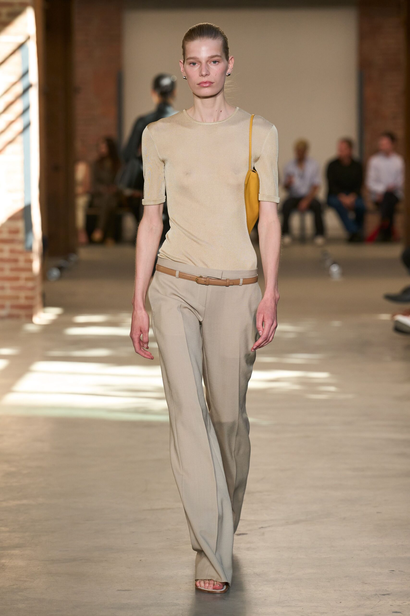 Sheer beige fine-gauge knit, elbow sleeves rolled into tiny cuffs. Mustard leather baguette tucked tight under the arm. Slim tan leather belt with a small oval buckle. Stone straight-leg trousers breaking softly over the sandal. Flat square-toe sandals. Walking through a quiet museum wing after the school groups leave.