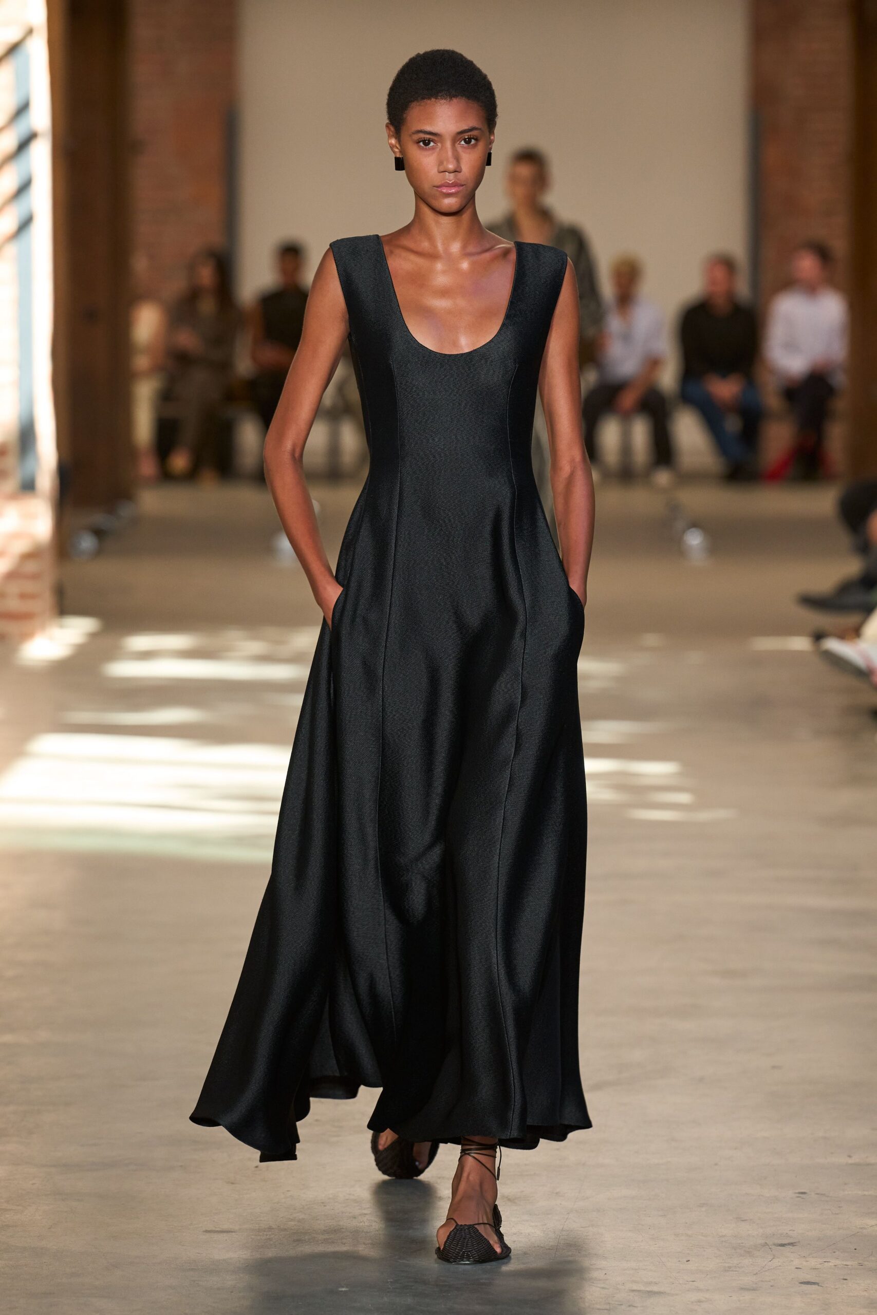 Small black rectangular studs. Black floor-length gown in dense satin, scoop neck and hidden side pockets. Flat thong sandals with woven black uppers and wraparound ties. Crossing a museum courtyard just before the string quartet starts.