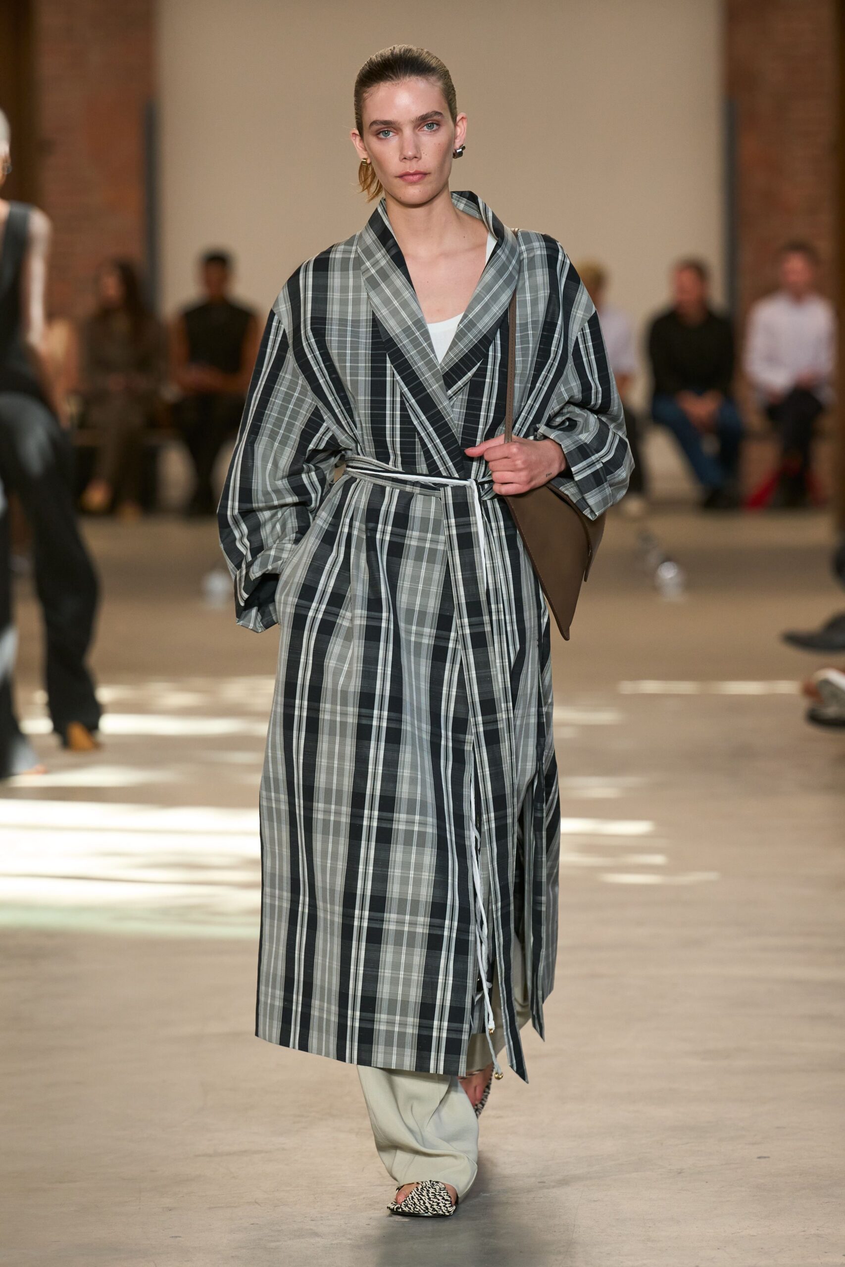 Grey-and-black plaid wrap coat with a shawl collar and cord tie ends. White scoop-neck underlayer, just visible at the chest. Pale dove trousers falling long over the foot. Flat brown shoulder bag tucked under one arm. Black-and-white woven pointed flats. Walking into a courthouse annex with coffee cooling in your hand.