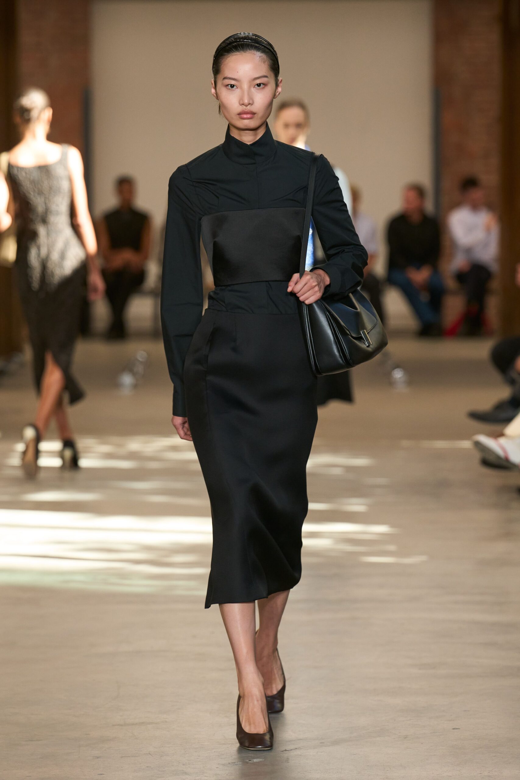Glossy black band holding the hair flat off the face. Black column dress with a satin bust panel over a stand collar. Soft black leather shoulder bag, folded body and slim silver tip. Dark square-toe pumps. For crossing a museum atrium before anyone else has arrived.
