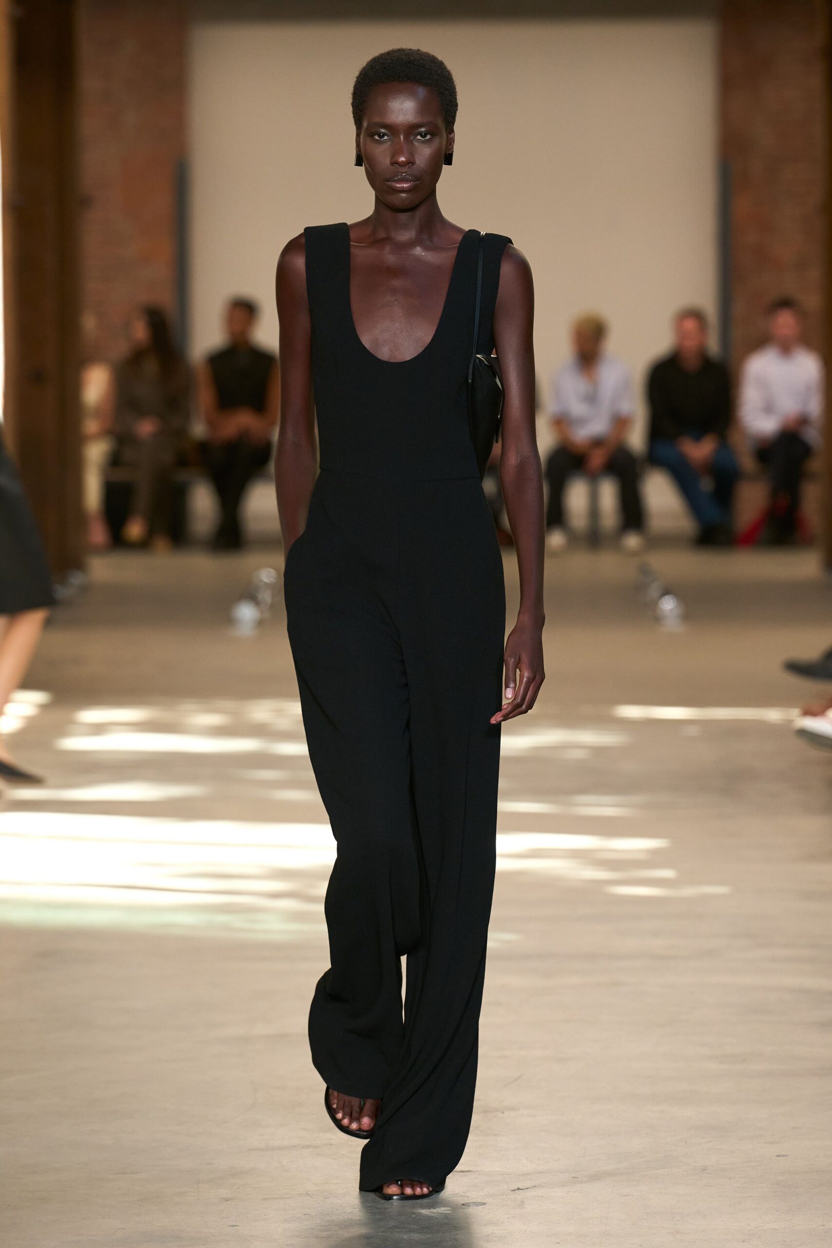 Black crepe jumpsuit with a deep scoop neck and pooled hems. Small black shoulder bag tucked tight under one arm. Barely there black sandals, square toes peeking past the hem. Glossy black block earrings. Crossing a museum atrium while the marble still holds the day's heat.