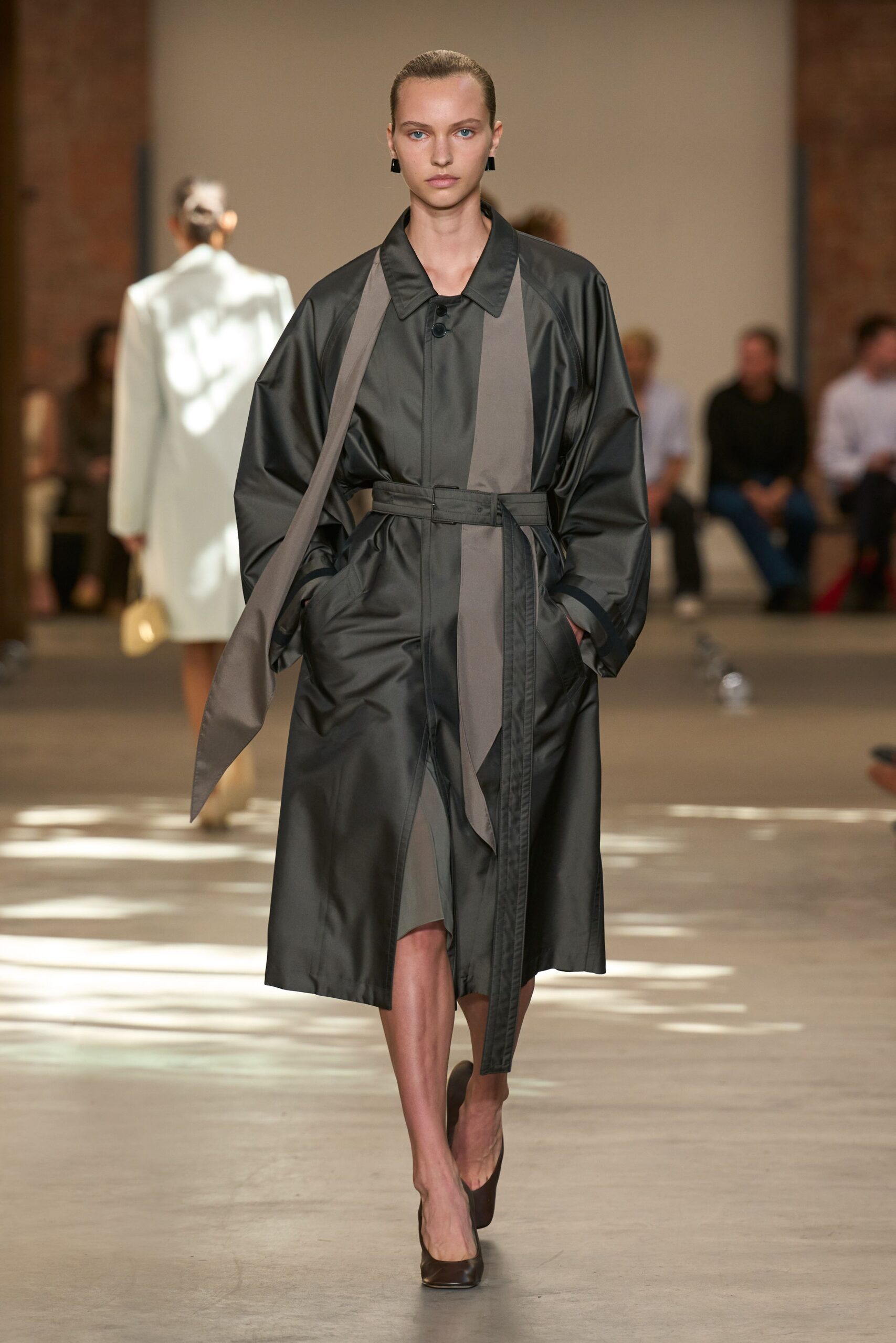 Charcoal satin-finish coat dress, cut with a storm flap hanging loose. Self belt threaded through a rectangular tab, one end dropping long. Small black block earrings. Dark brown pumps with a blunt squared toe. Crossing an office plaza just after rain, carrying news nobody wants.